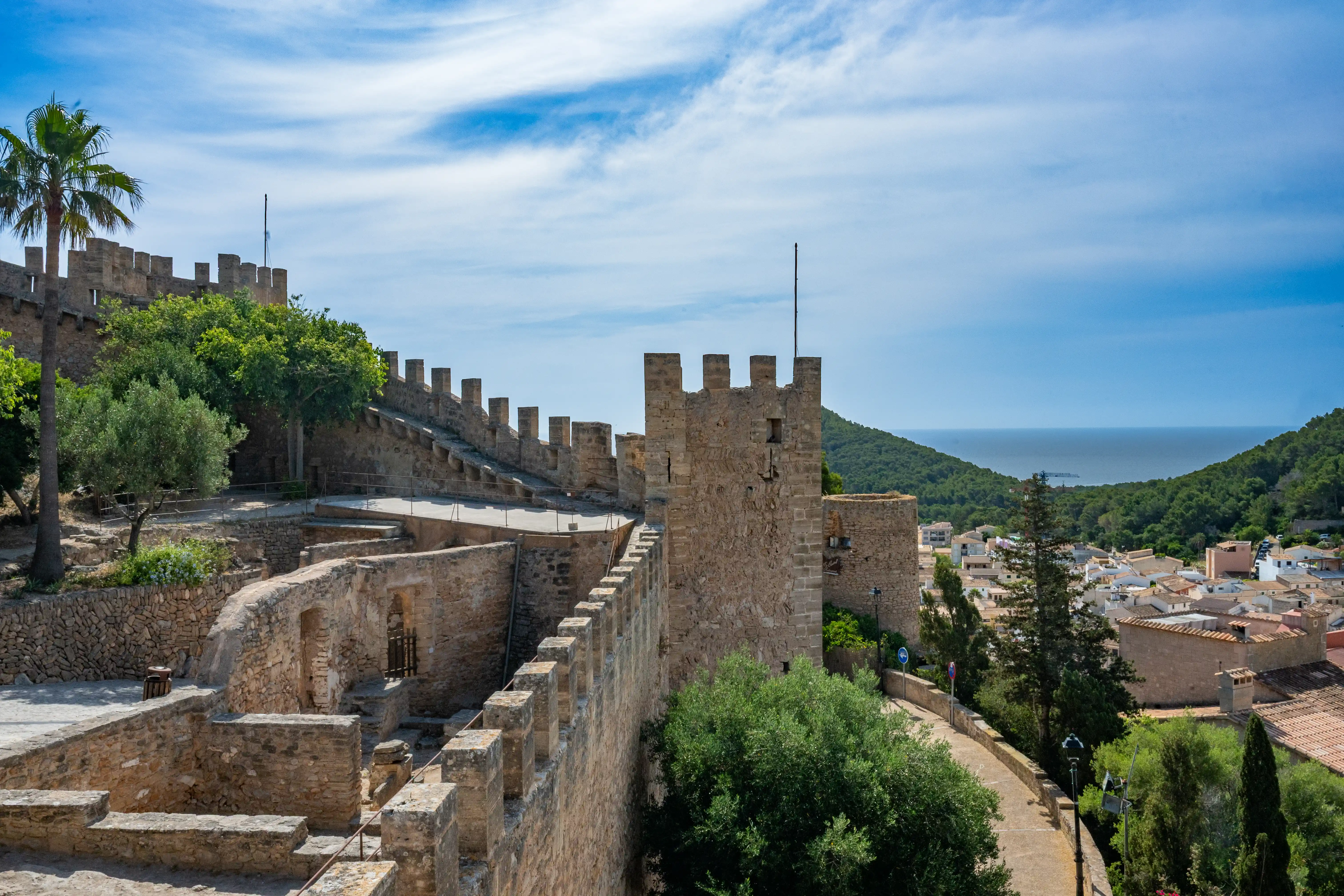 View from a historic stone castle wall with crenellations, looking down onto a coastal village with white buildings and red-tiled roofs, surrounded by green hills and leading to the blue sea under a sky with white clouds.