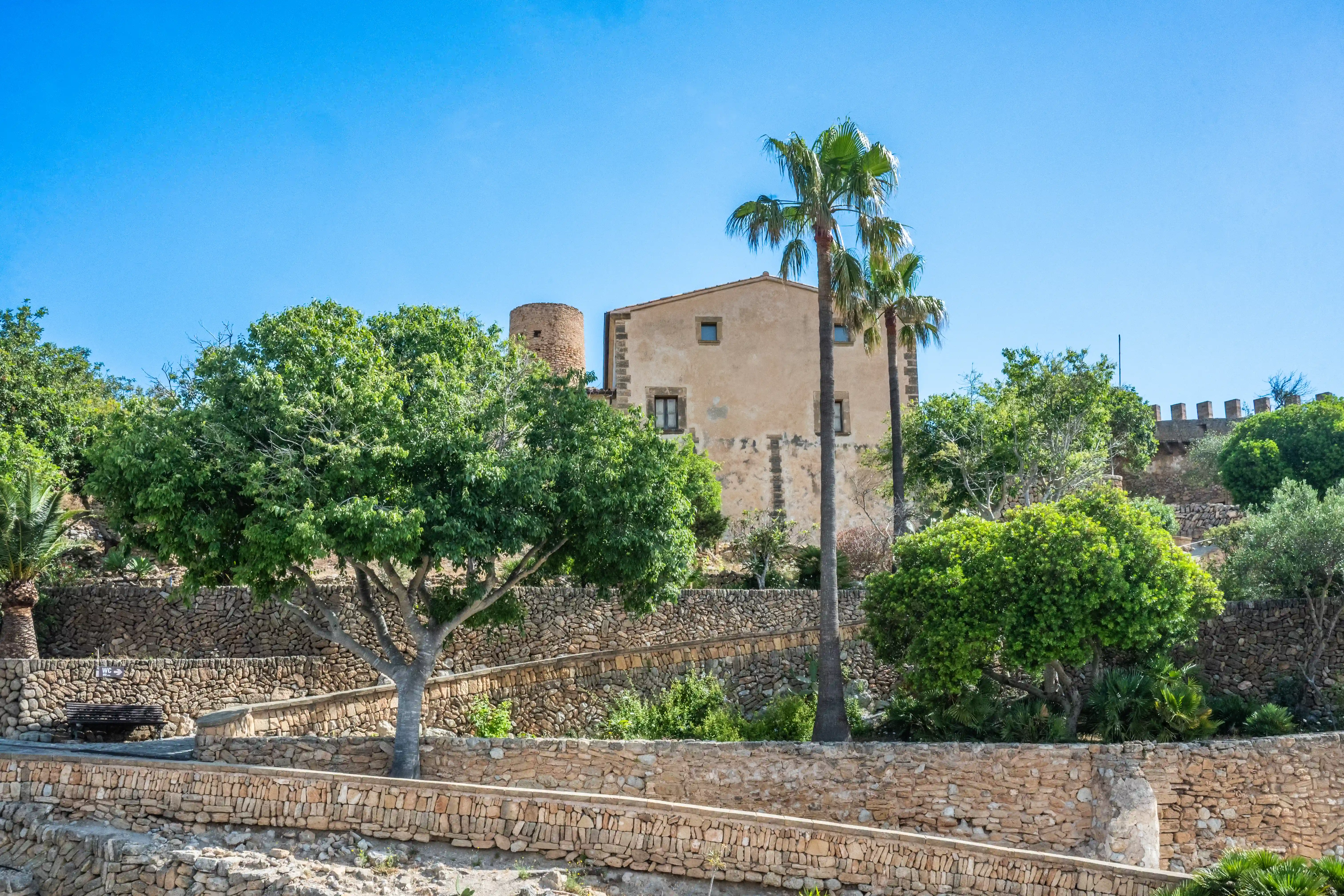 A view of an old stone tower and a beige building with windows, surrounded by green trees and palm trees, with stepped dry-stone walls in the foreground, under a bright blue sky.