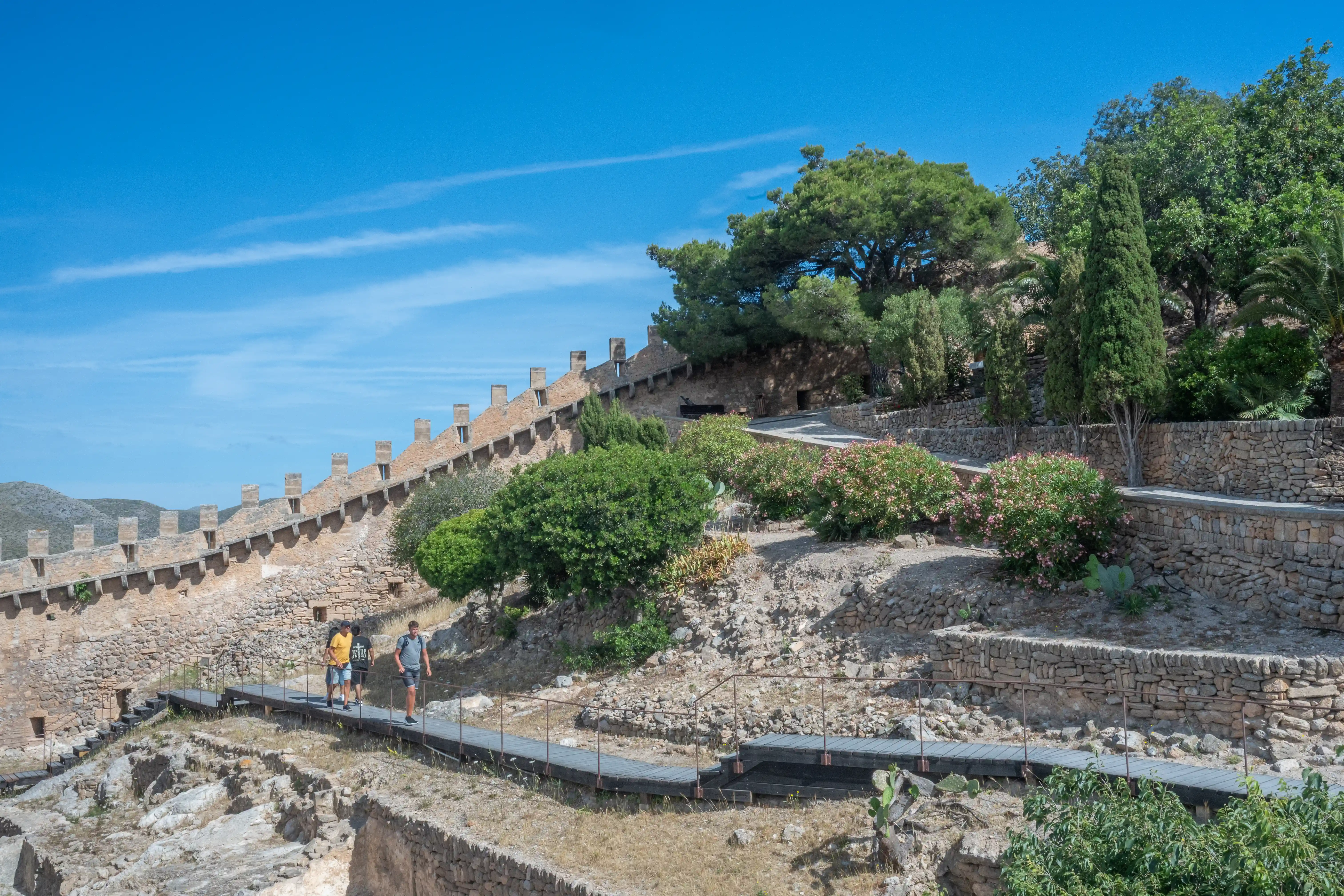 Three people walk along a wooden pathway in front of a weathered stone fortress wall on a sunny day with a blue sky.