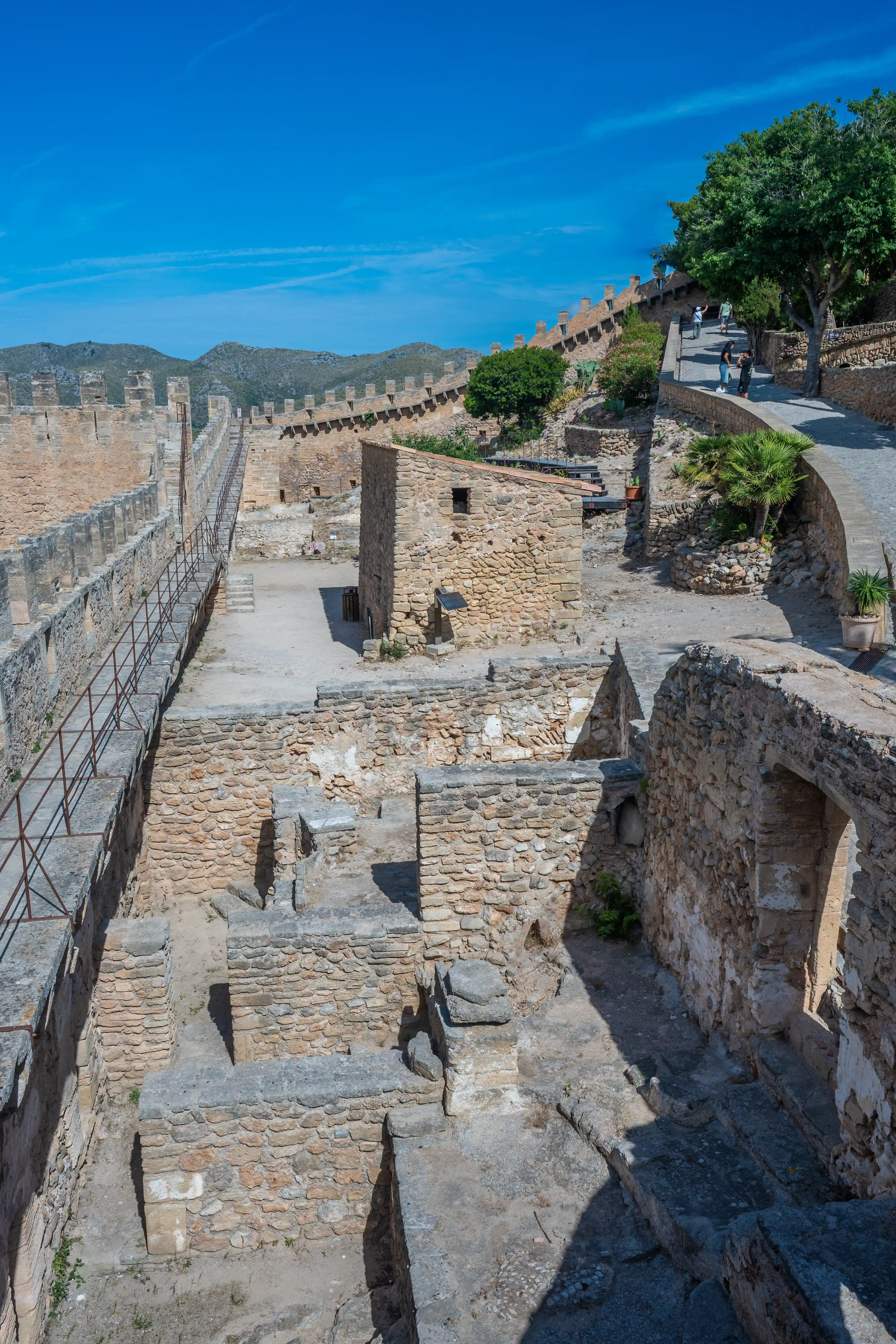 Wide shot of a historical fortress, showcasing its weathered stone walls, ramparts with crenellations, and interior ruins. A paved pathway lined with trees and potted plants runs along the top of the wall, with several tourists walking on it. In the background, arid hills rise against a vibrant blue sky.