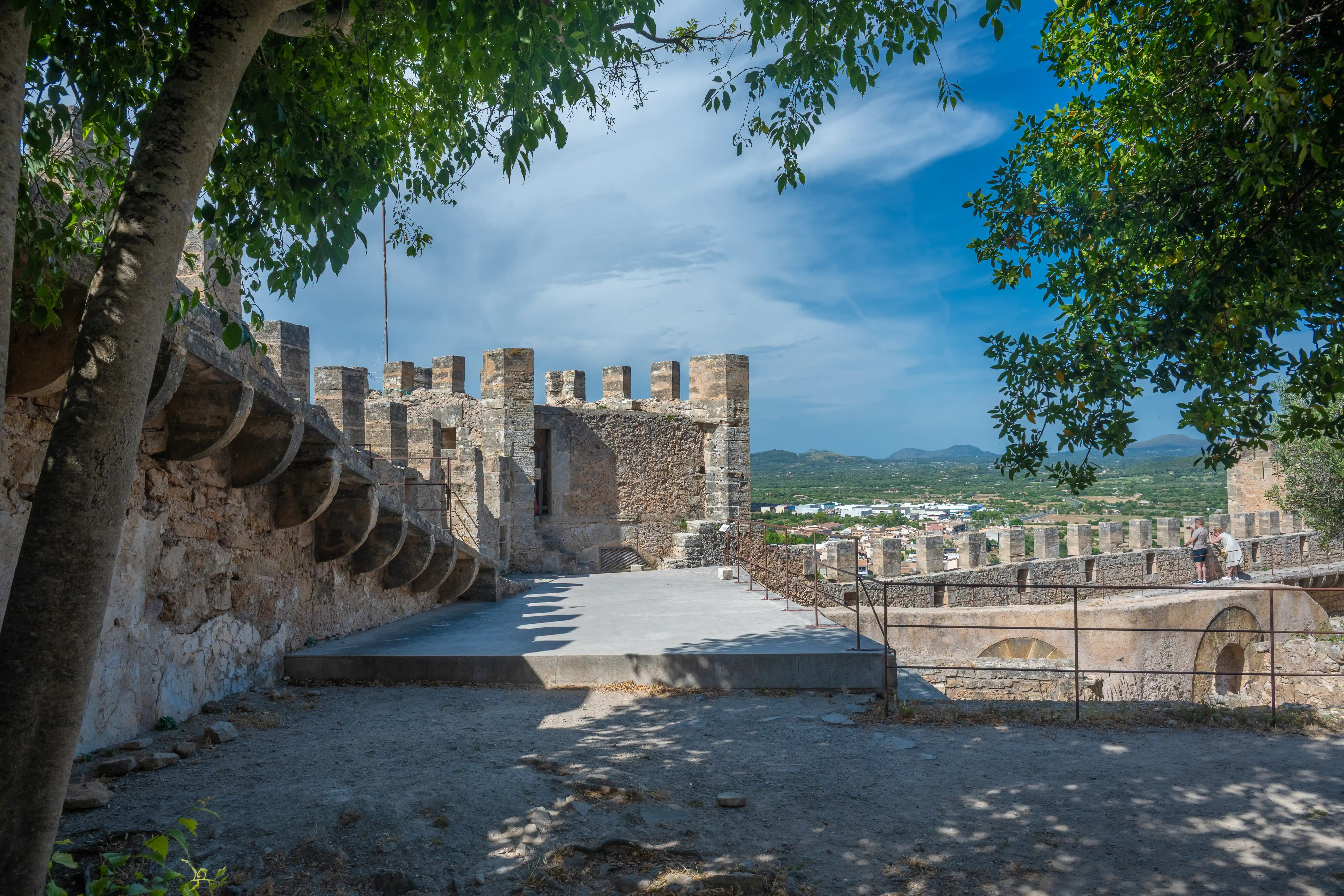 Stone battlements of an old castle with a town and hills in the distance, framed by green tree branches and dappled sunlight.