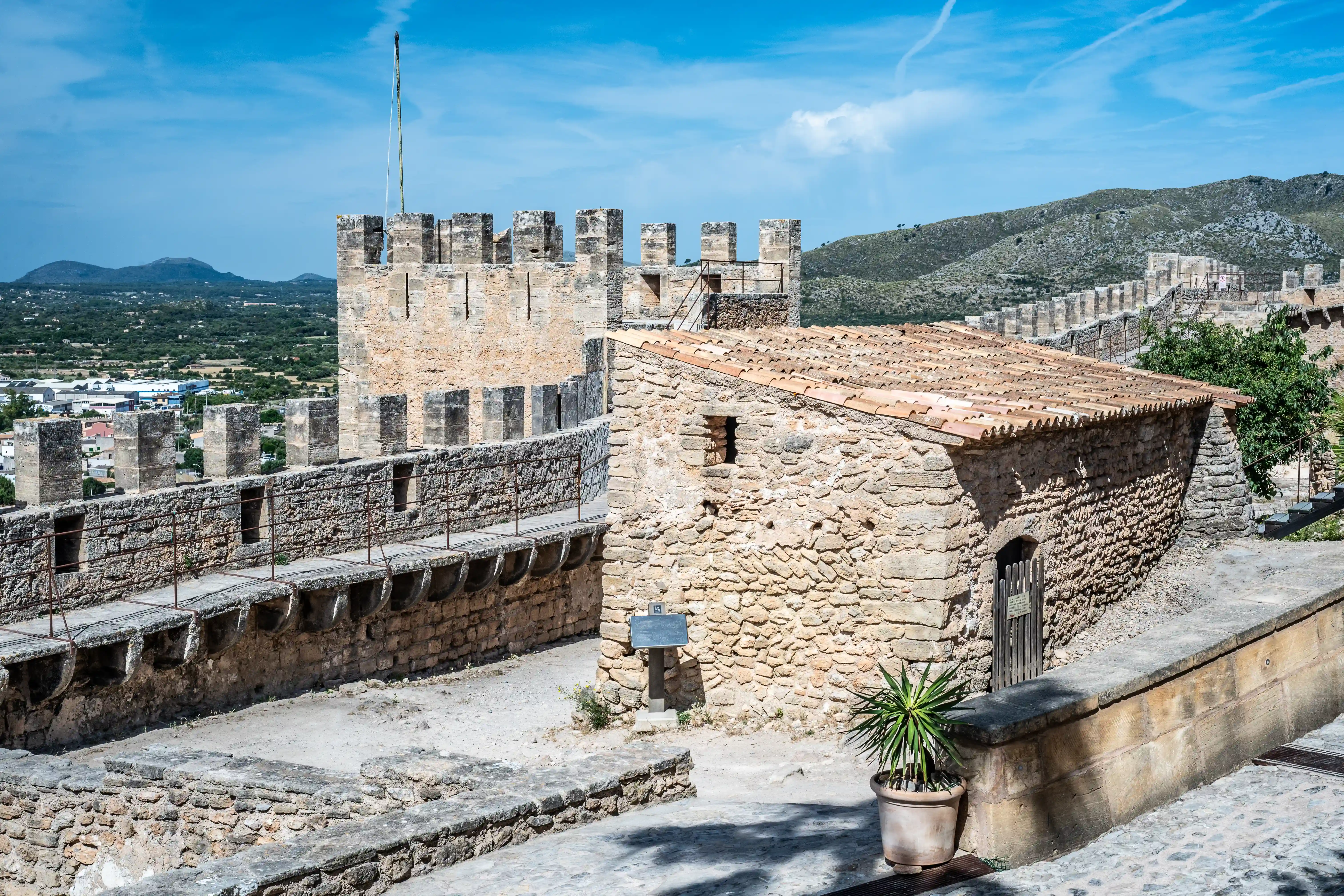 A wide shot of a historic stone castle with battlements and a small attached building with a tiled roof, set against a backdrop of green hills and a blue sky with scattered clouds.