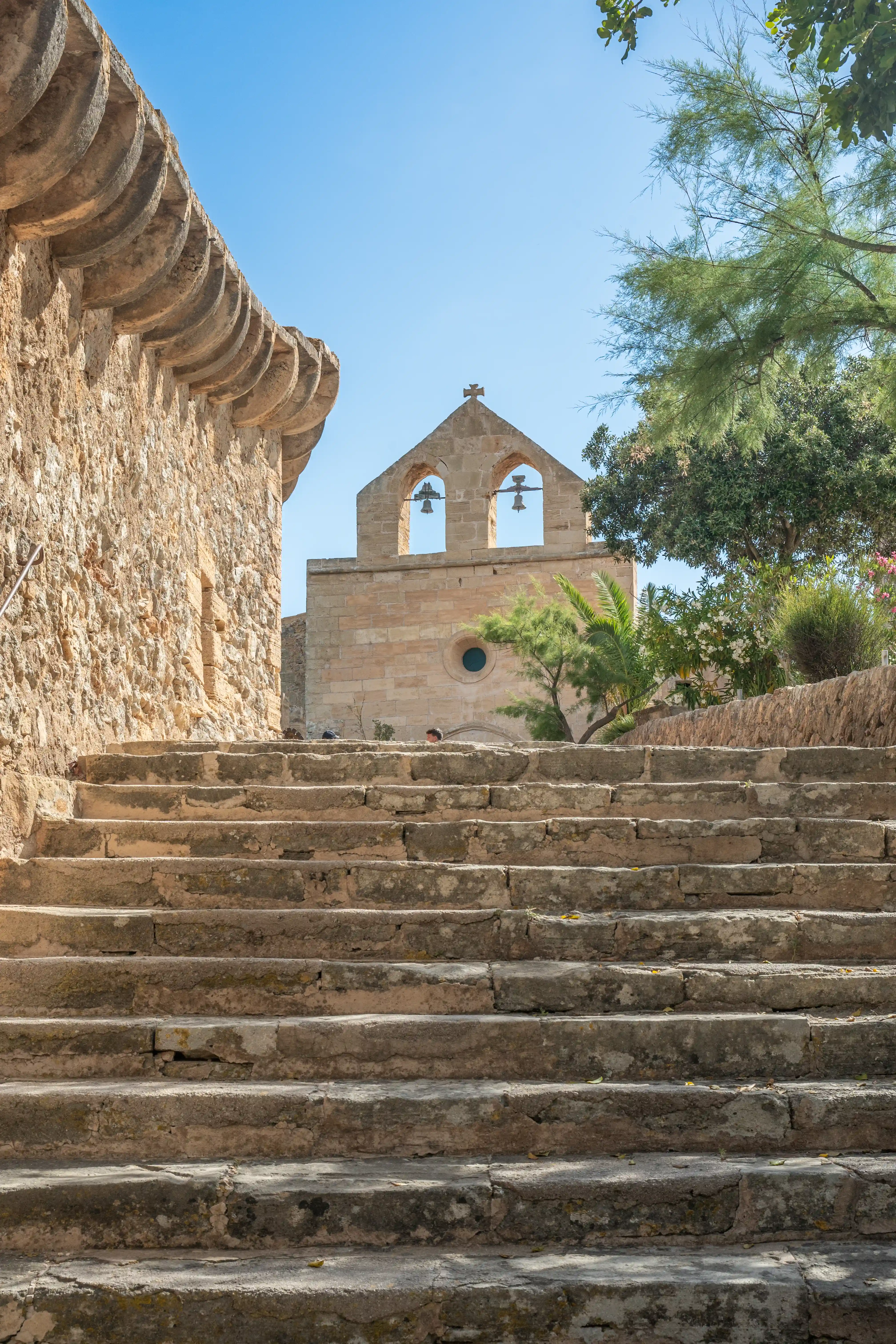 A rustic stone staircase ascends towards a charming bell tower with two bells, set against a vibrant blue sky, bordered by an ancient stone wall and verdant foliage.