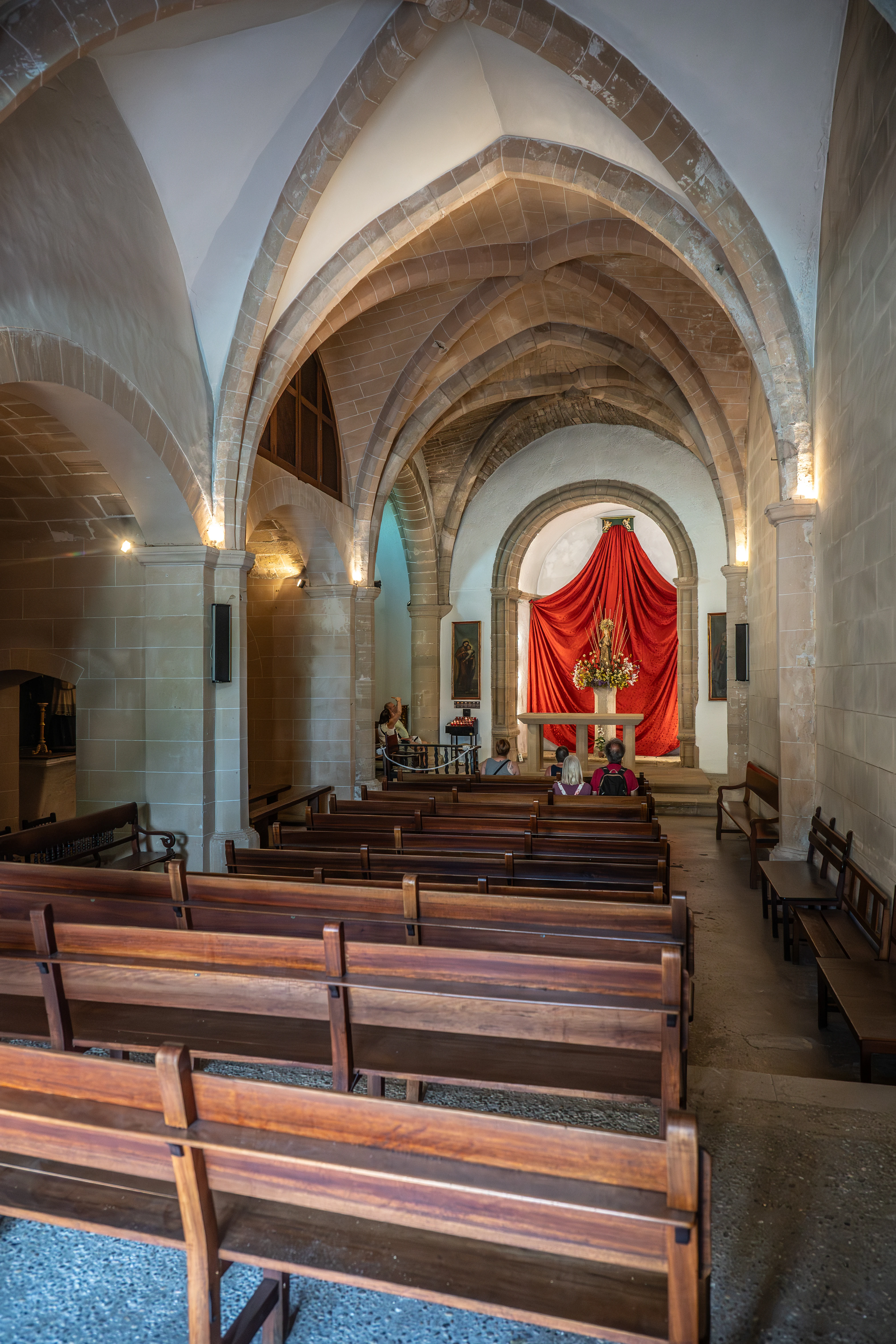 Interior of a stone church with arched vaulted ceilings, leading to an altar draped in red fabric. Rows of empty wooden pews fill the foreground and middle ground, with a few people visible seated further back.