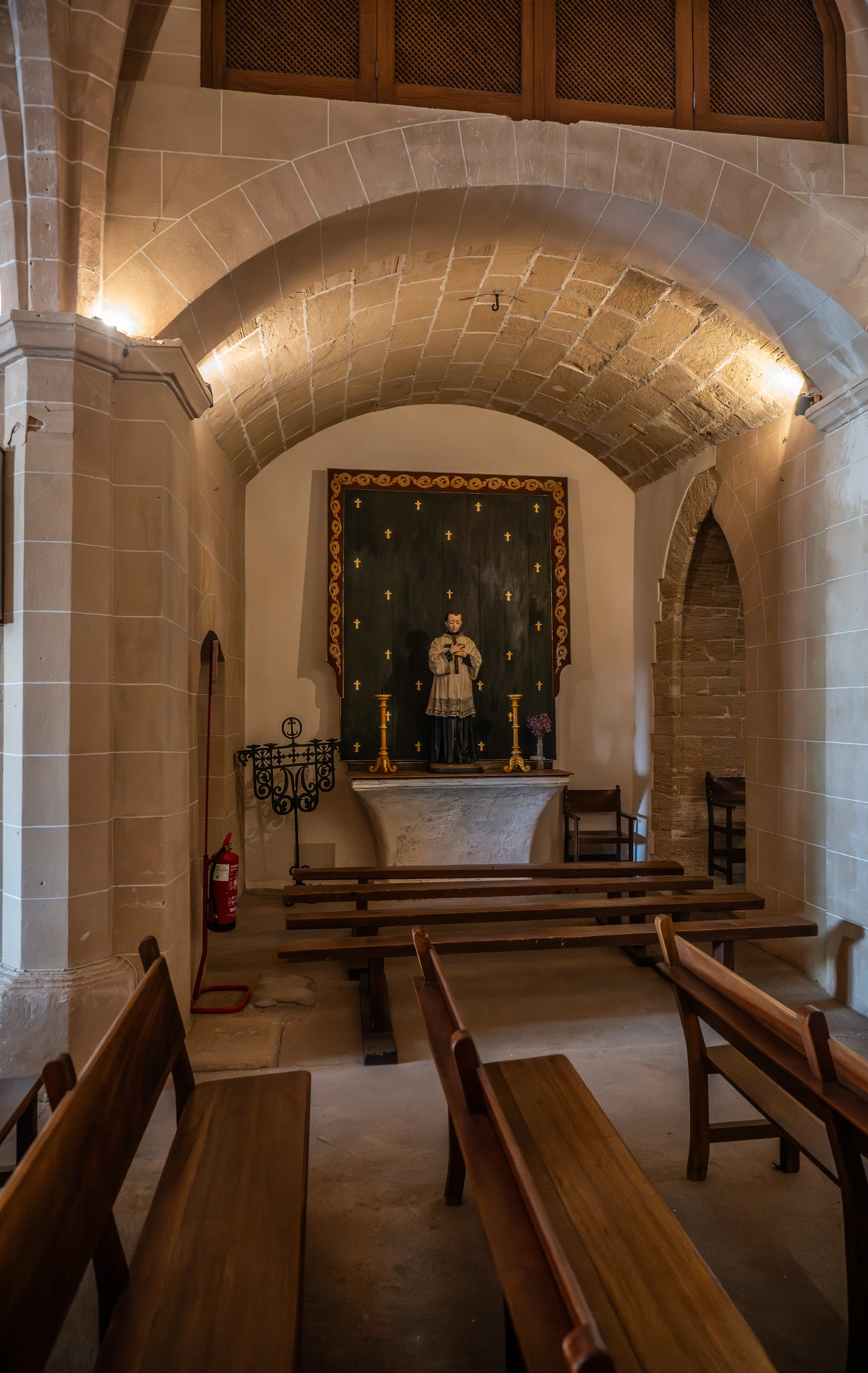 A view of the inside of a chapel showing a statue of a saint on an altar, surrounded by wooden pews and stone architecture.