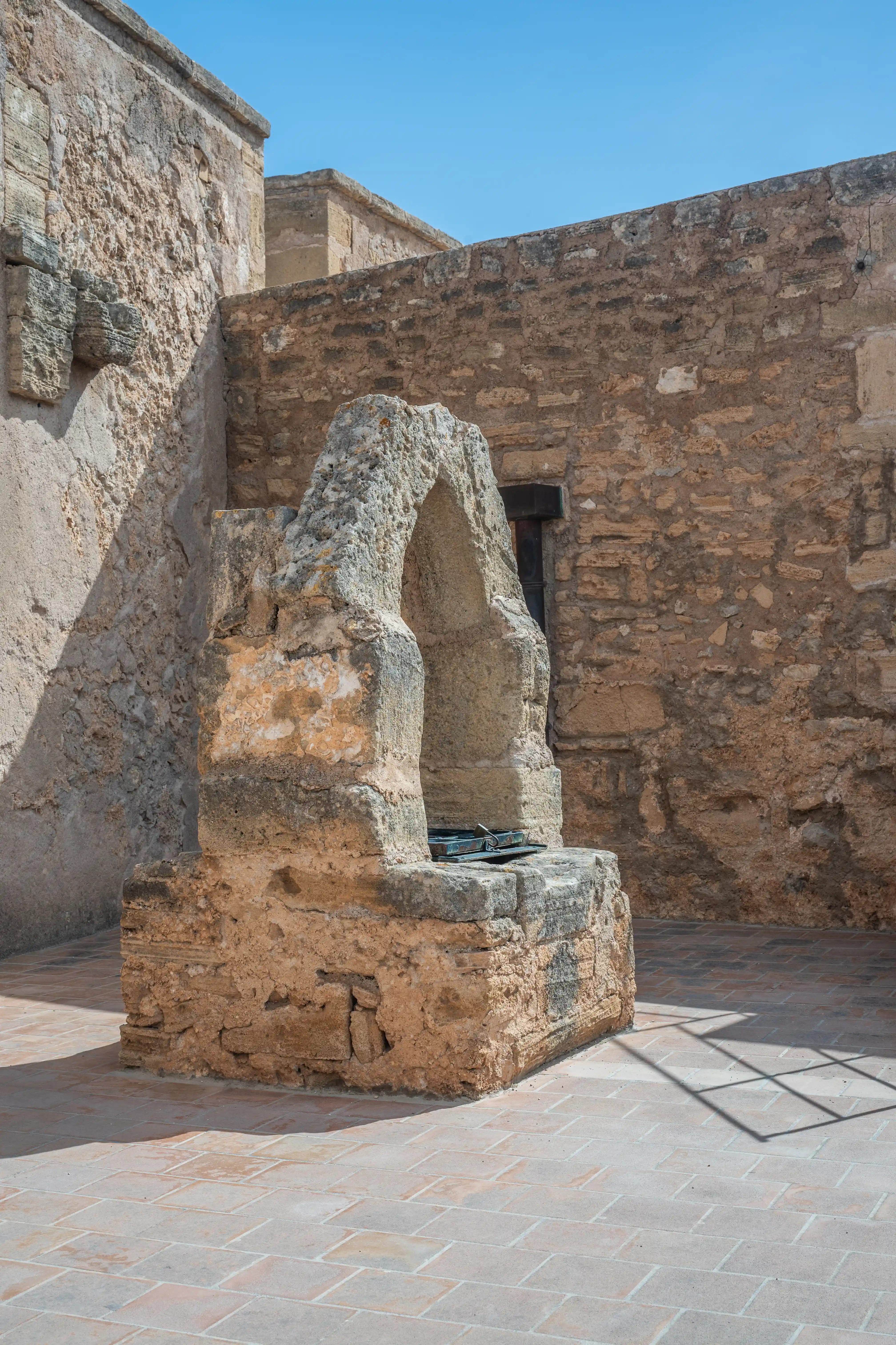 An old stone well with a rough arch stands in a paved courtyard with sandstone walls under a clear blue sky.