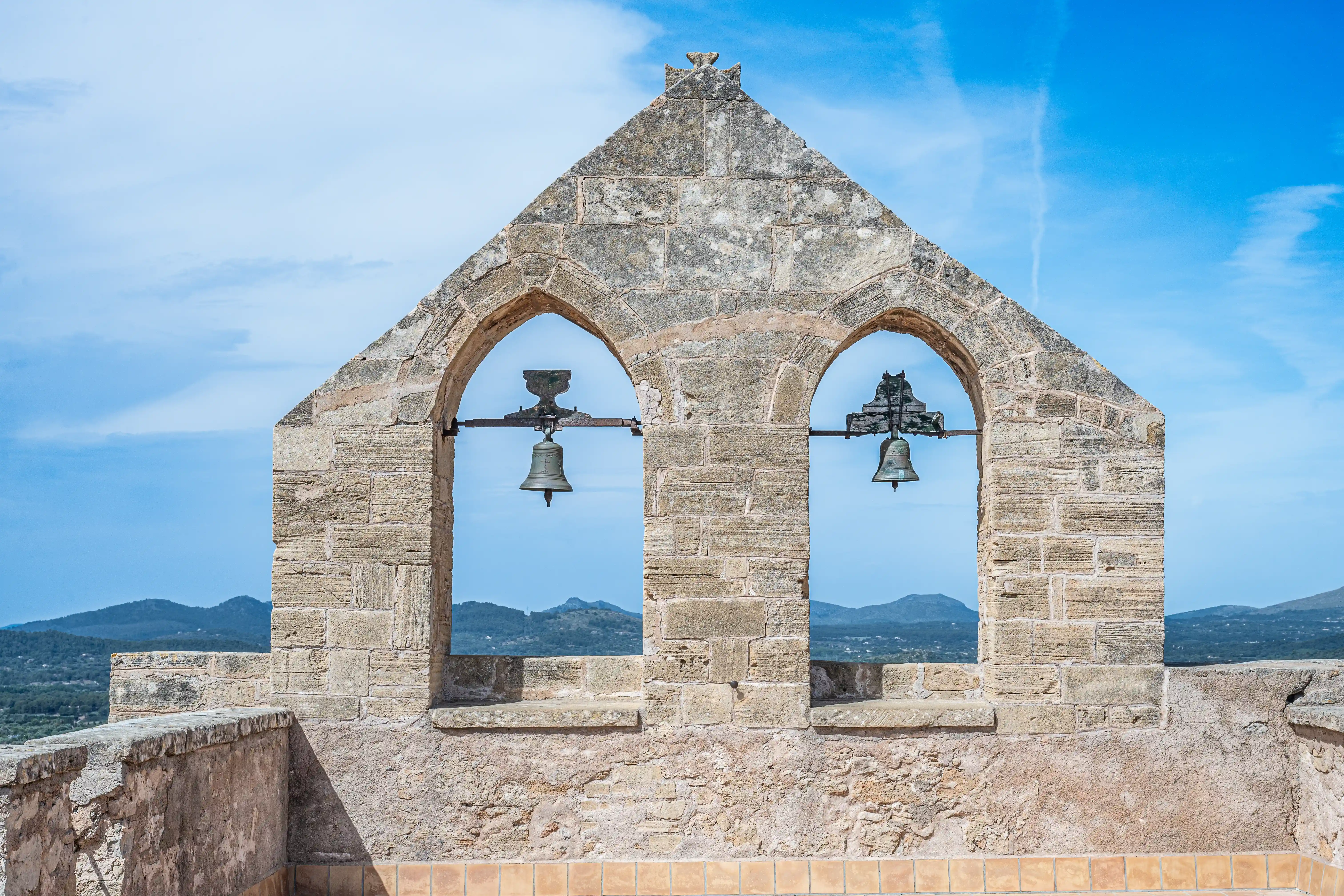 A stone bell tower with two bells visible through arched openings, set against a bright blue sky and distant green hills.