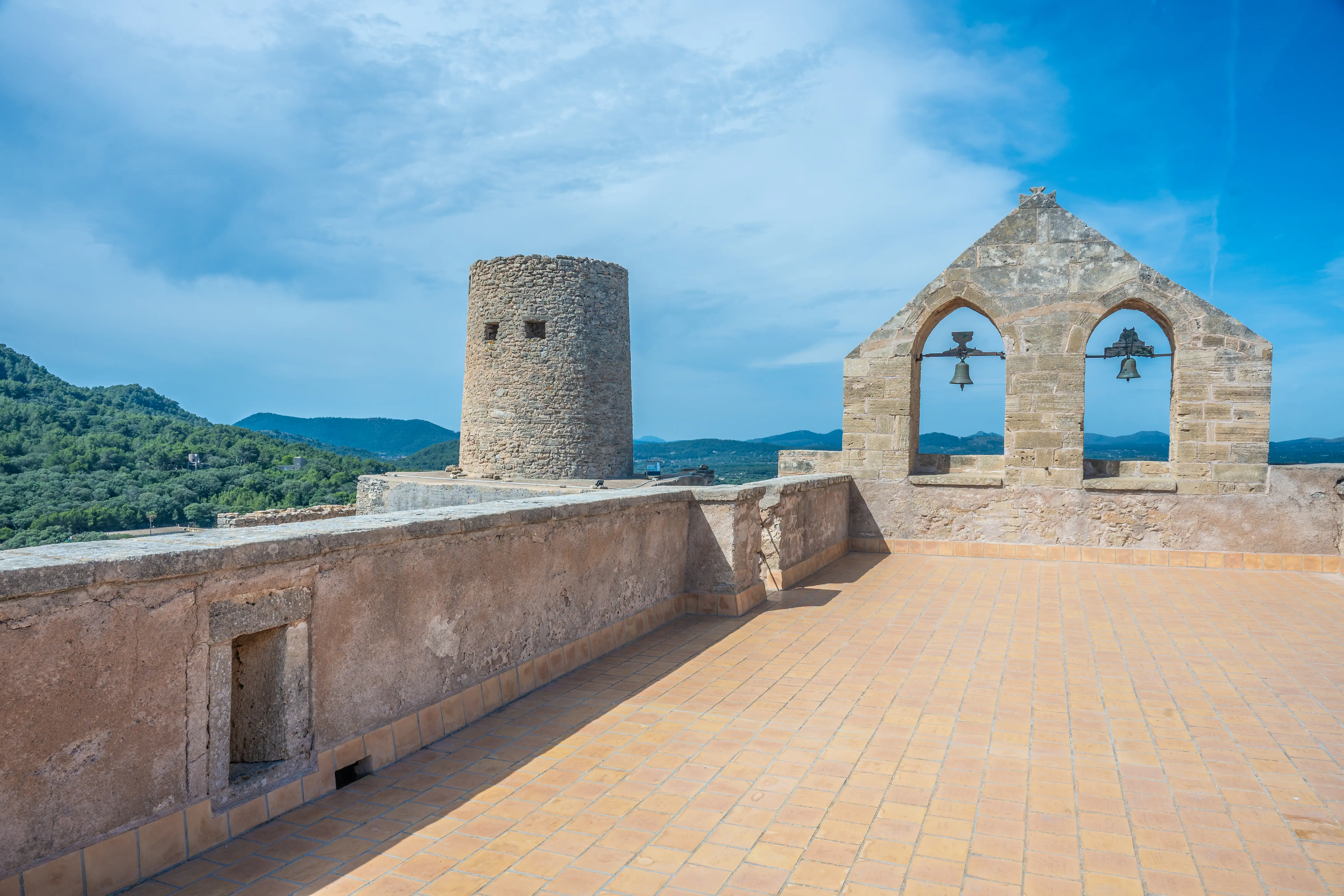 A wide shot of a historic stone fortress's rooftop, showing a terracotta tiled floor, a stone balustrade, a round stone tower, and an arched belfry with two bells. In the background, rolling green hills and mountains are visible under a clear blue sky.