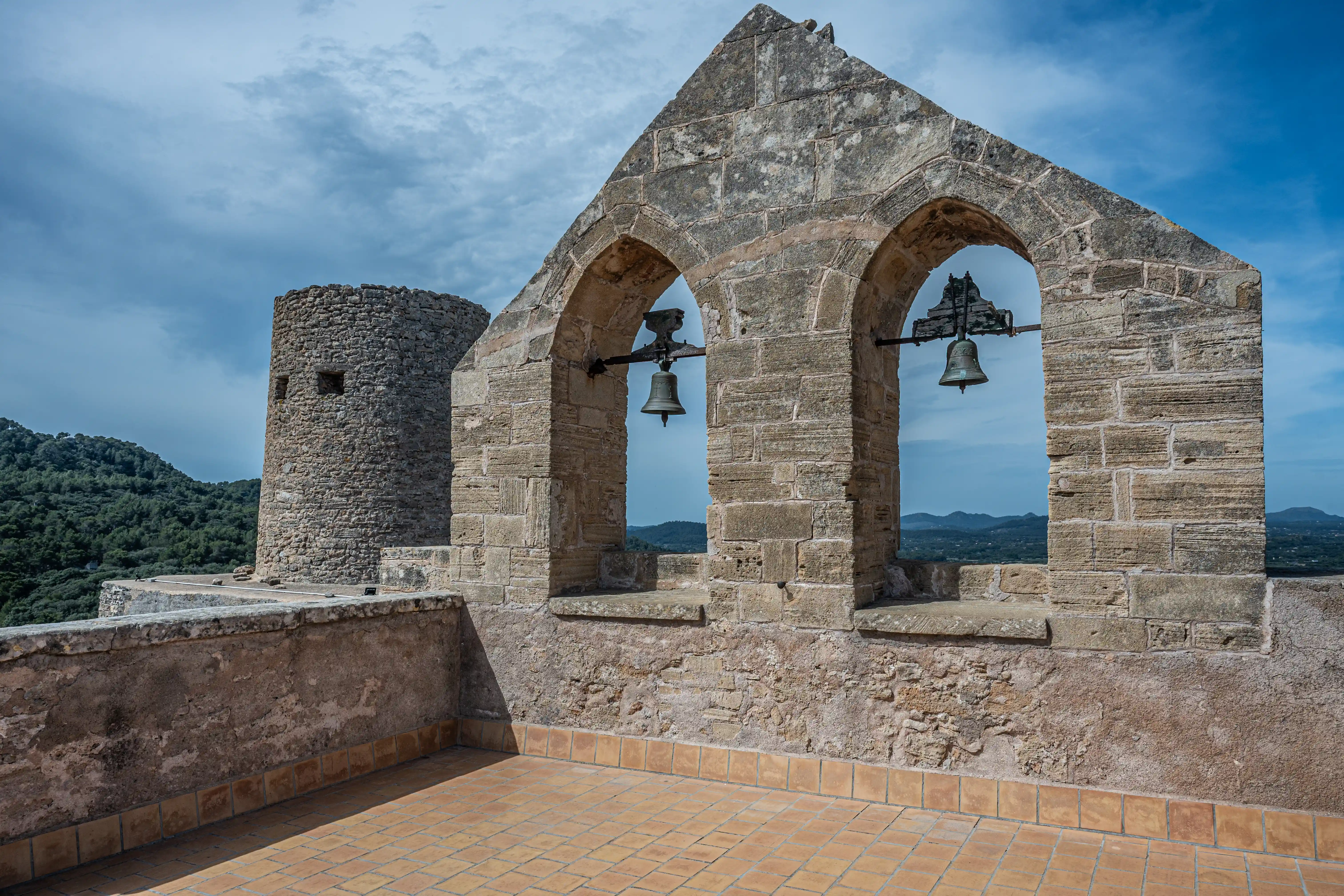 View from a historic stone terrace featuring a bell tower, archway with bells, and a landscape of hills under a cloudy sky.