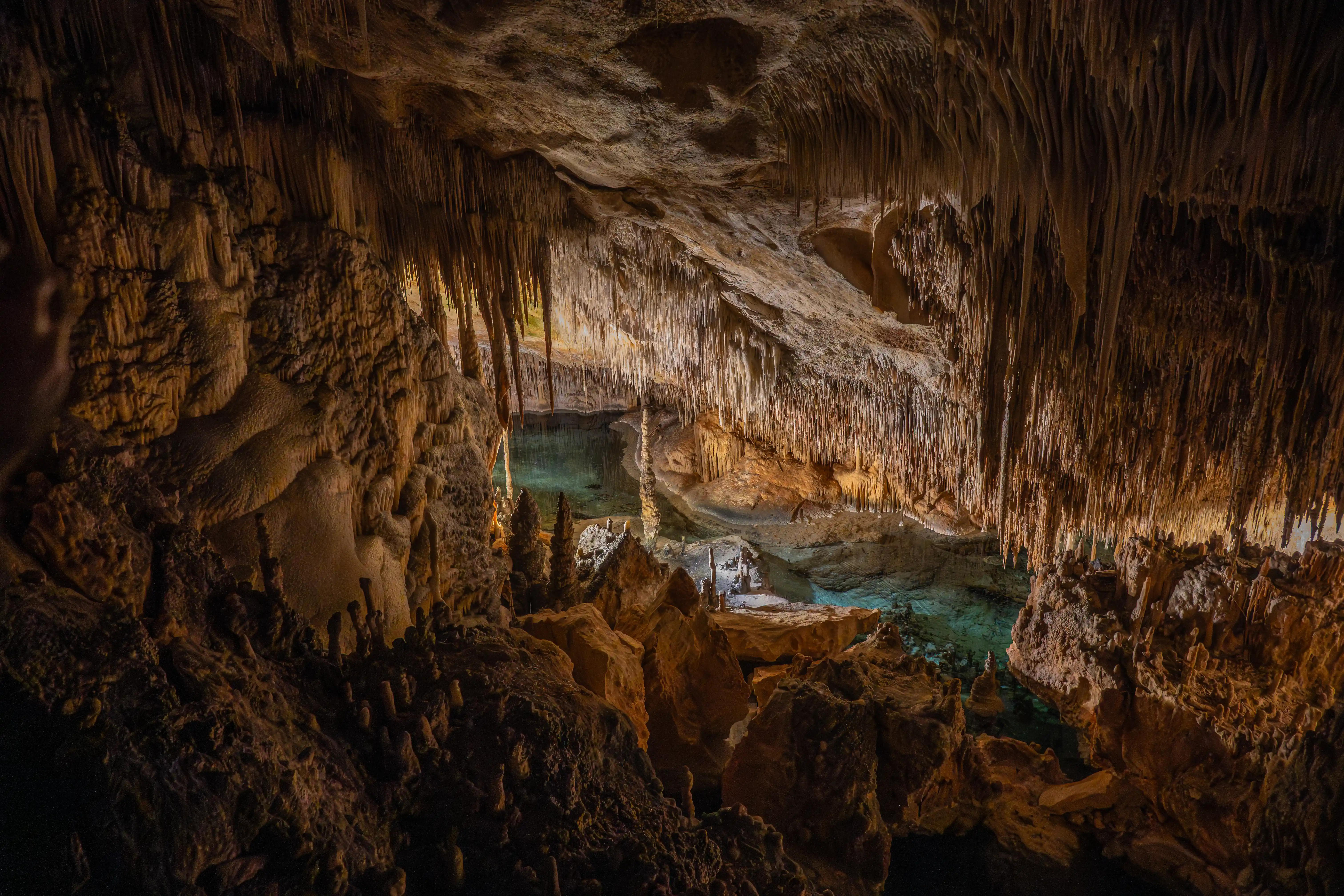 A dimly lit cave interior with numerous stalactites and stalagmites, featuring a serene pool of turquoise water.