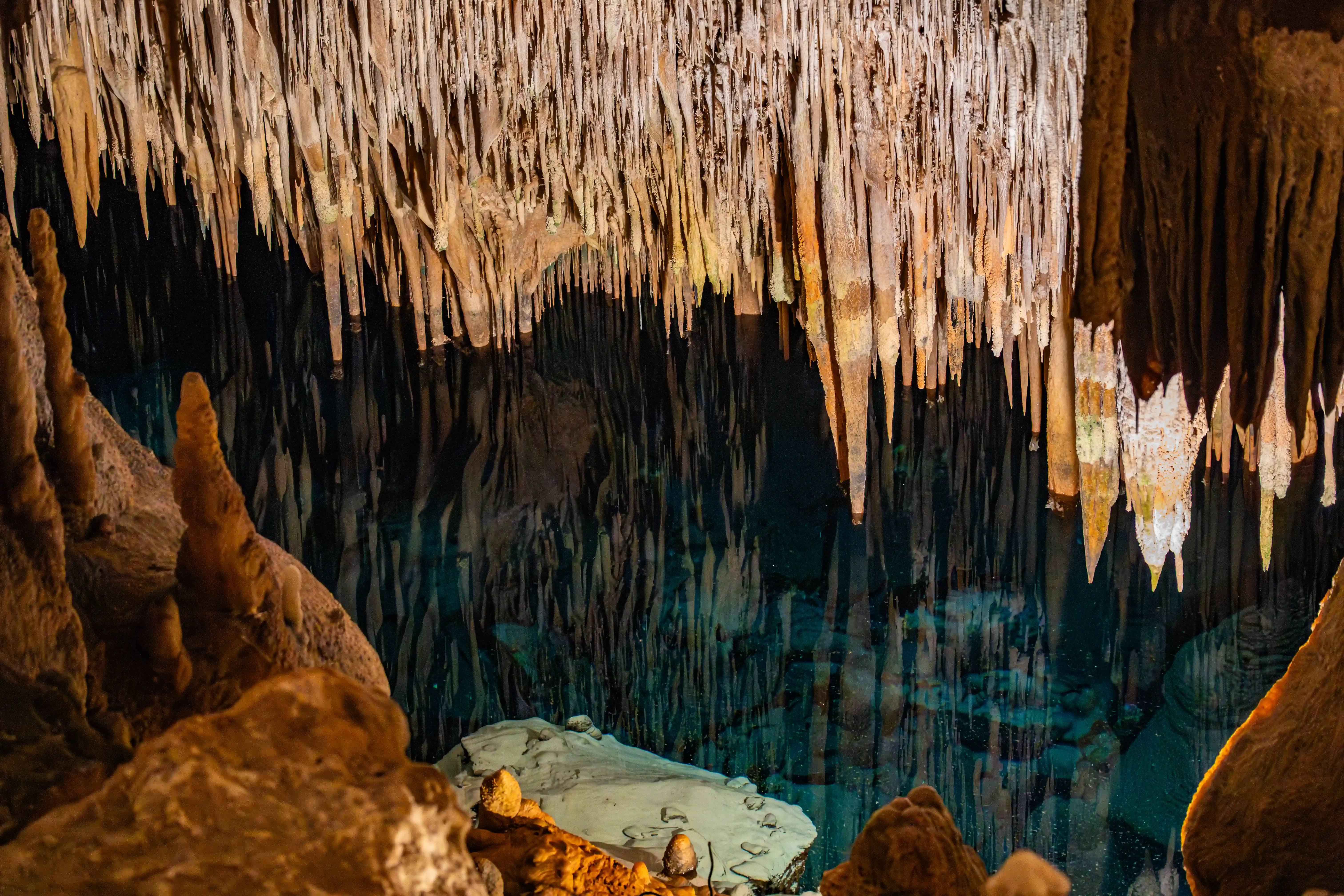 A cave interior with numerous beige and brown stalactites hanging over a body of clear, turquoise water. Submerged stalagmites and rocks are visible beneath the water's surface, with the cave walls reflected in the water.
