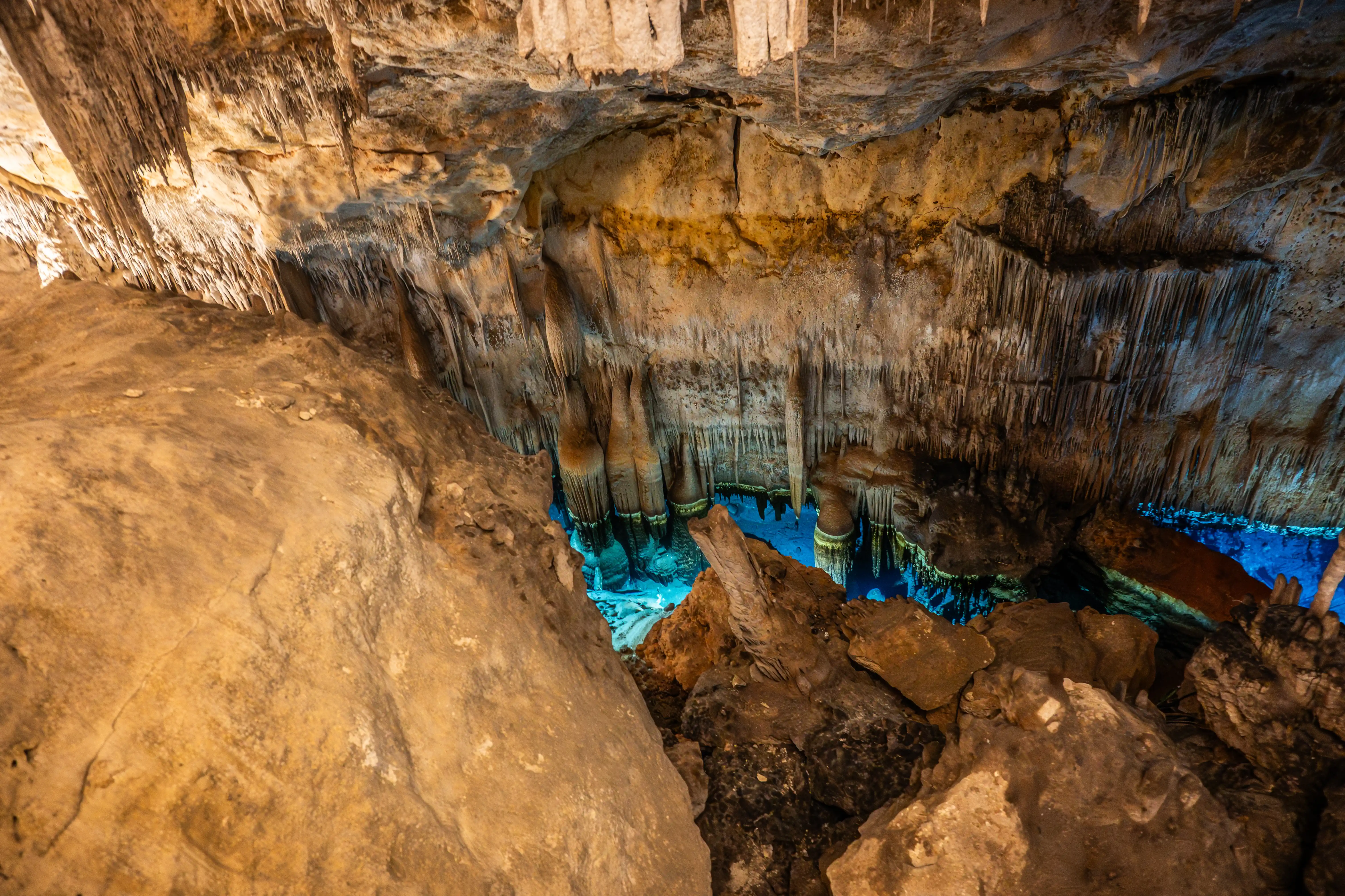 A cave interior with numerous stalactites hanging from the ceiling. In the centre of the image, a pool of vibrant blue water is visible, surrounded by rock formations and more stalactites. The lighting is warm and dramatic.