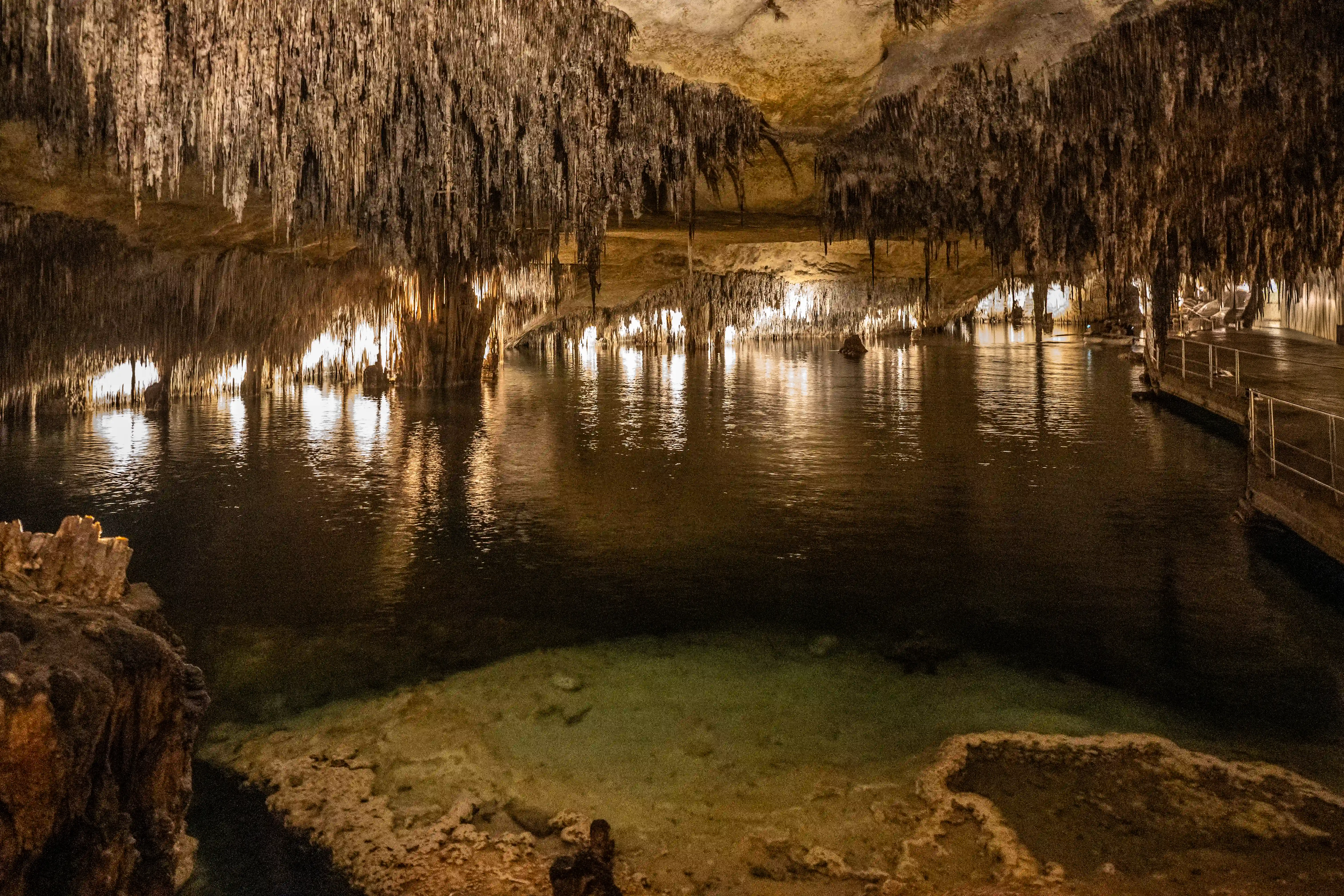 A subterranean cave chamber with a tranquil lake reflecting numerous stalactites hanging from the ceiling. A pathway with a railing runs along the right side of the cave, bordering the water.