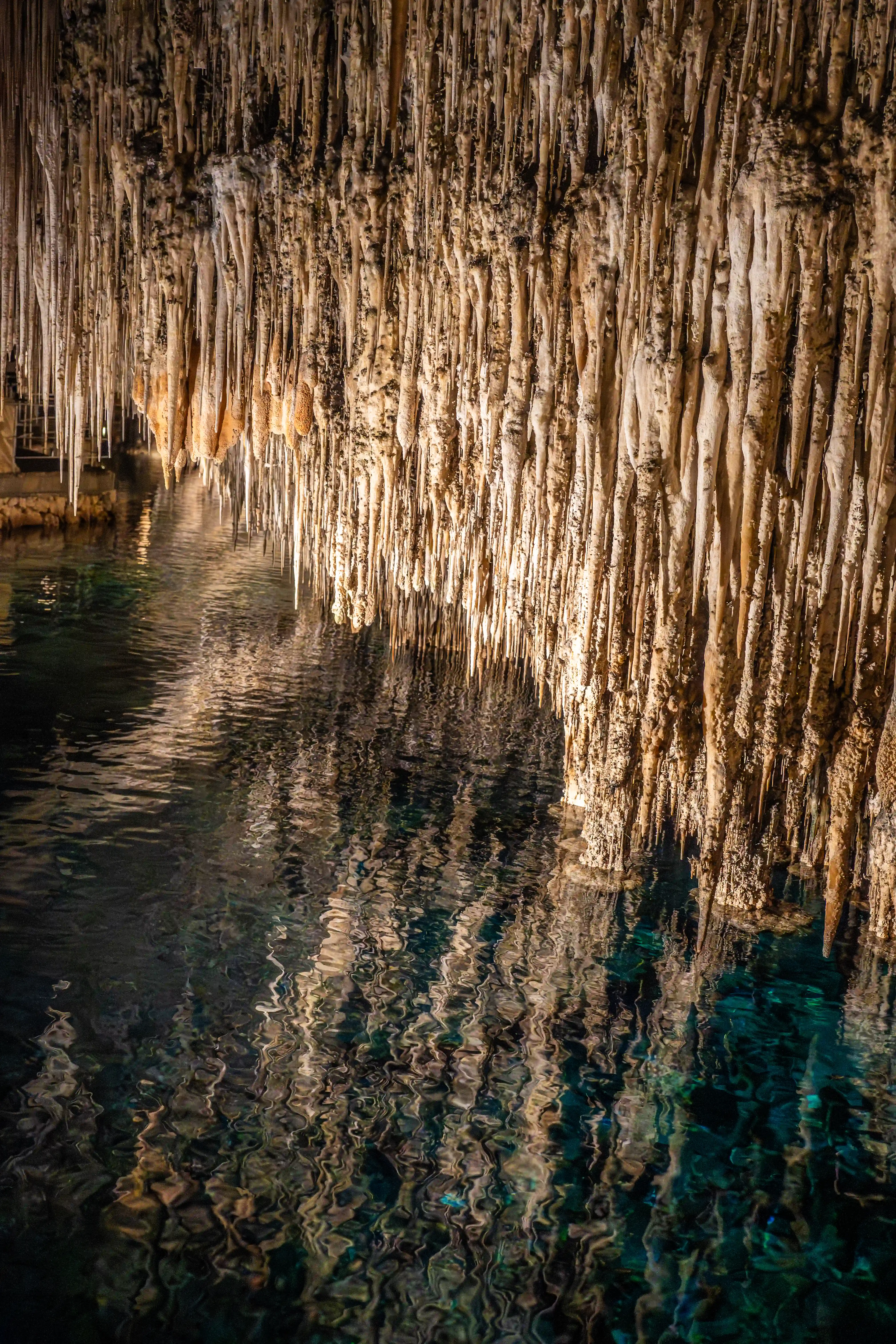 Numerous natural, cream-coloured stalactites hang from a cave ceiling, casting reflections on the dark, clear water below.