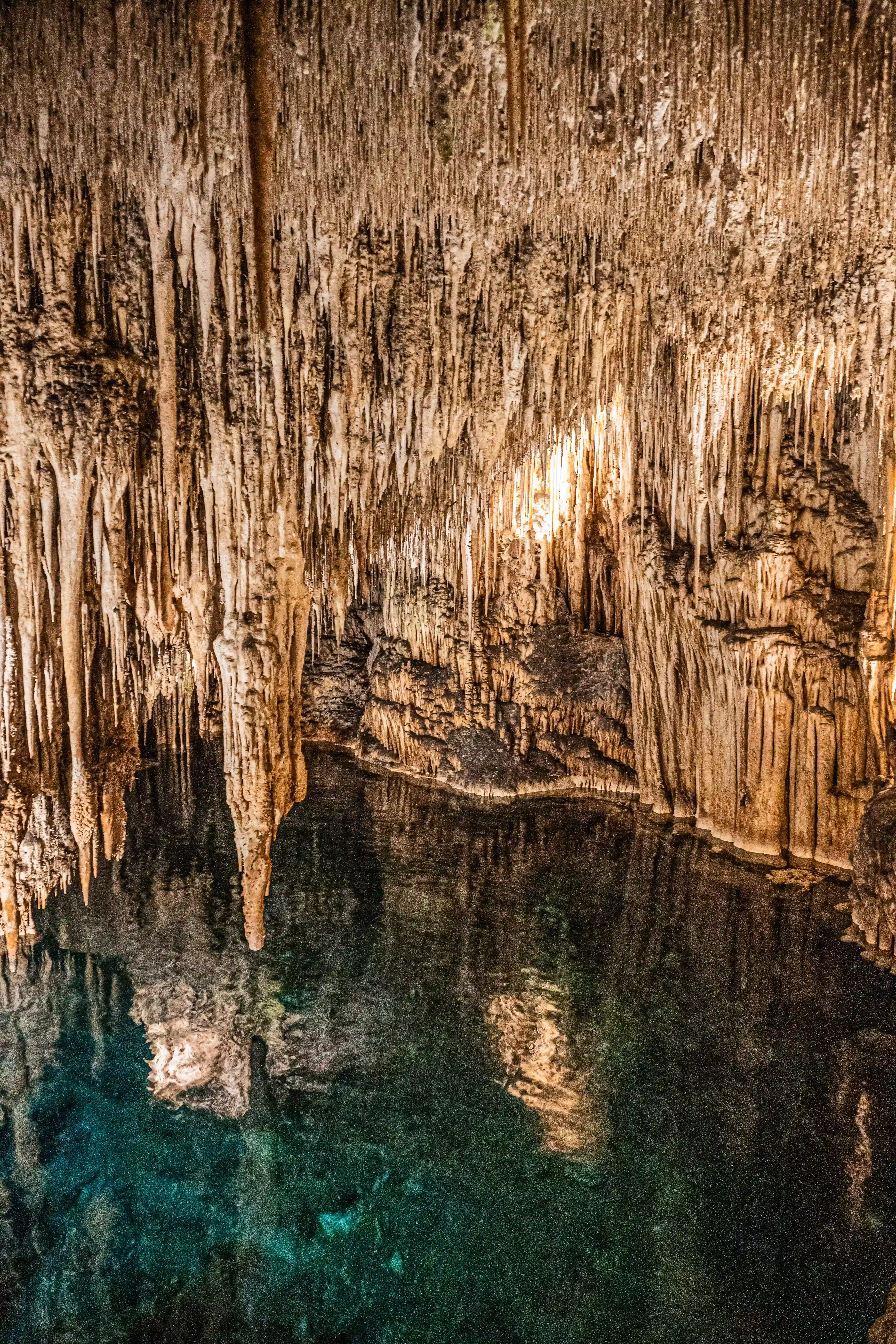 A photograph capturing a cave interior with numerous beige and brown stalactites hanging from the roof, overhanging a deep, clear turquoise pool of water that mirrors the formations above.