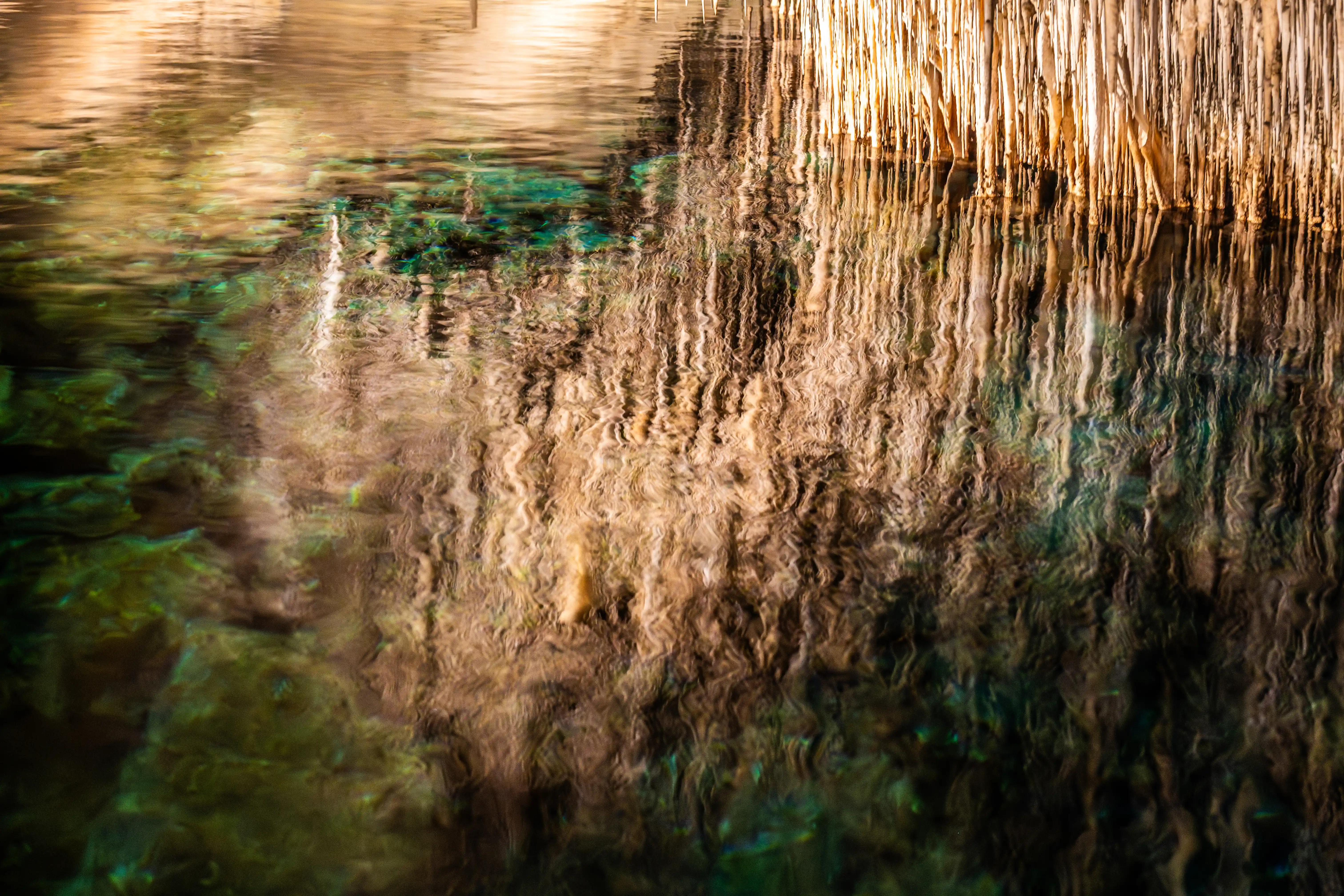 Abstract reflection of stalactites in rippling water, showing distorted vertical lines of brown and tan, with patches of dark green and blue.