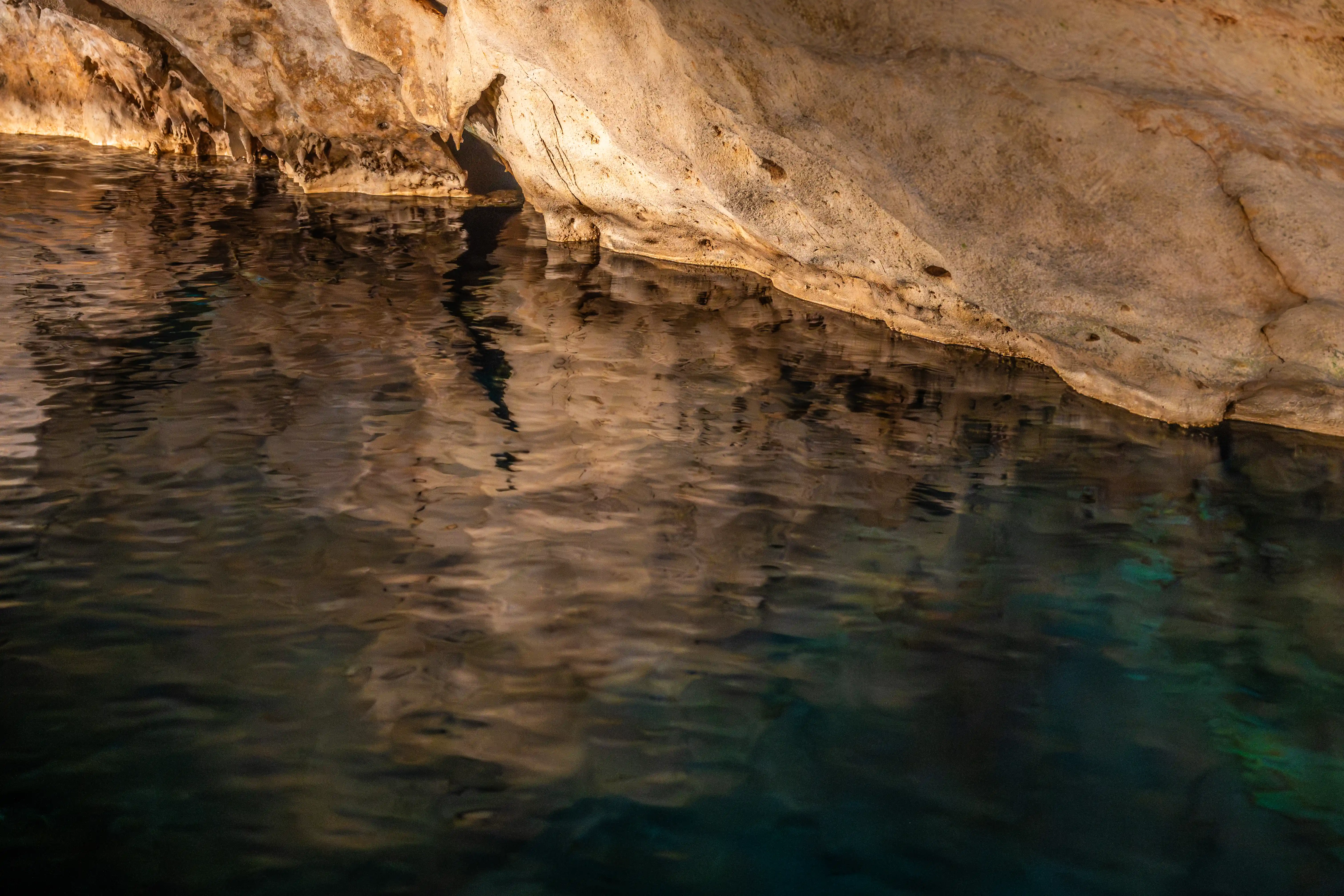 A close-up of a cave's interior, showing a still, dark pool of water with its surface reflecting the textured, light-coloured cave walls and ceiling.