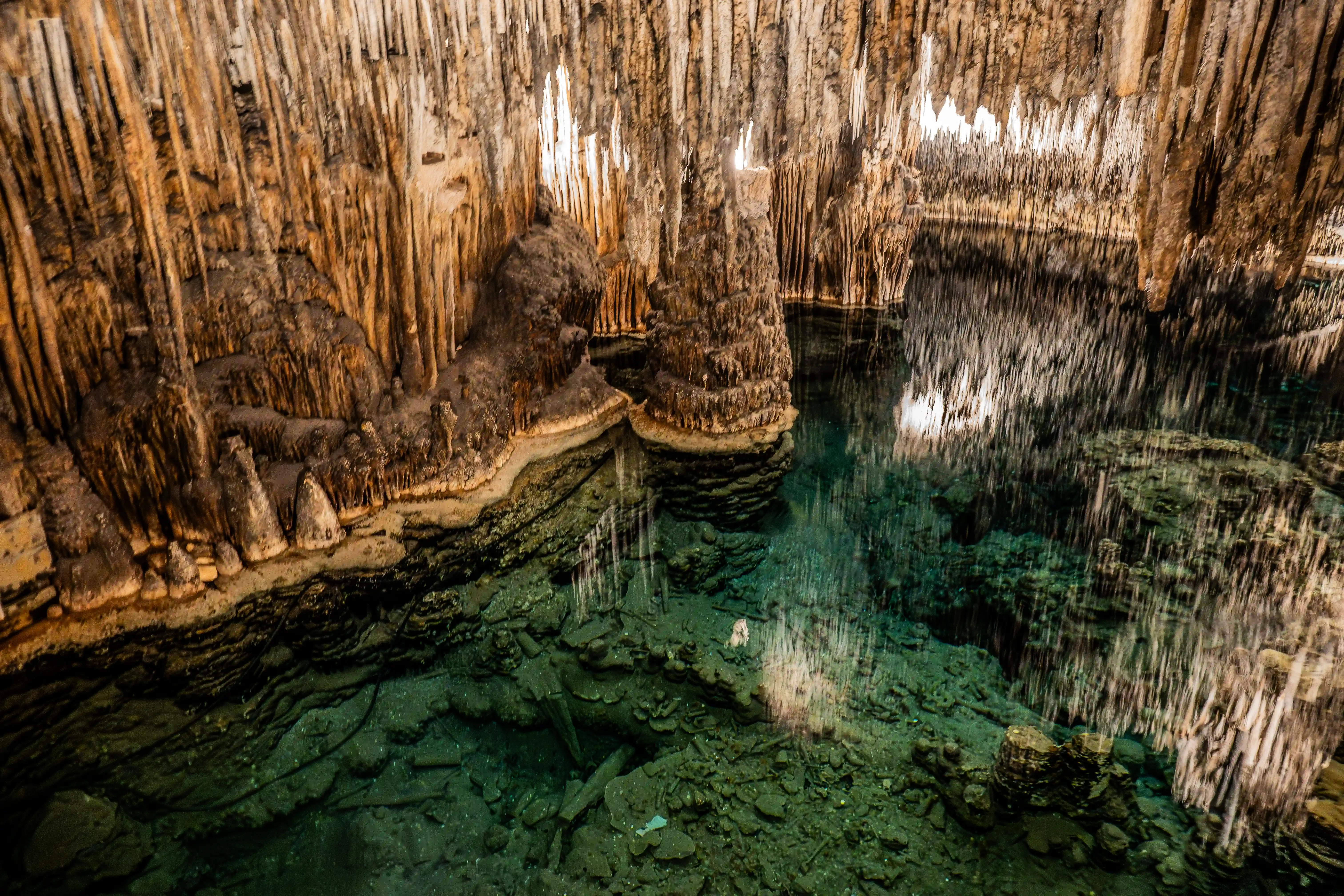 A cave interior with numerous stalactites hanging from the ceiling and stalagmites rising from the floor, surrounding a clear turquoise lake that reflects the formations.