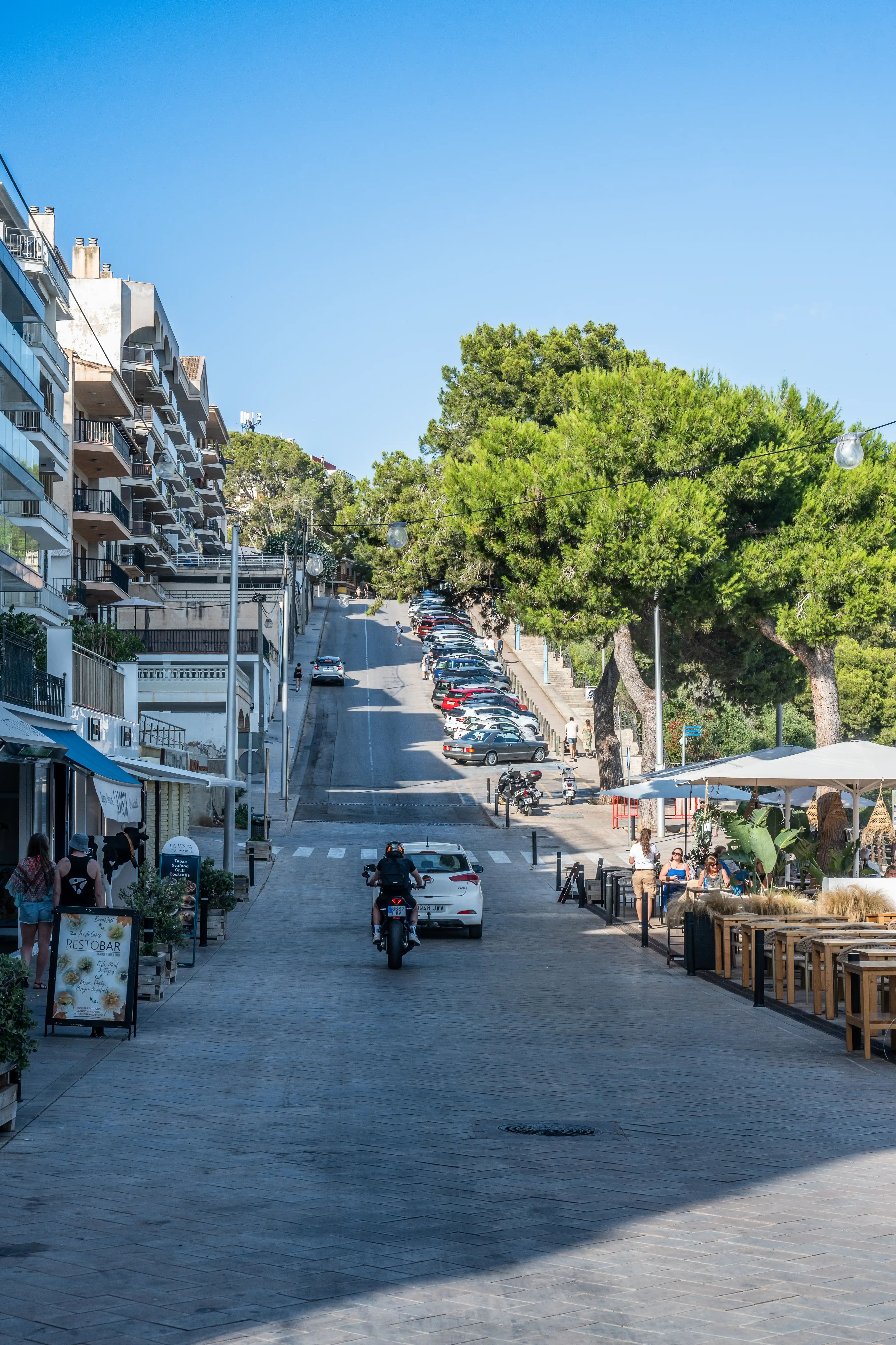 A paved street slopes uphill in a Mediterranean town, bordered by apartment buildings with balconies on the left and trees and an outdoor restaurant on the right. Cars are parked along the road, and a motorbike and pedestrians are visible.