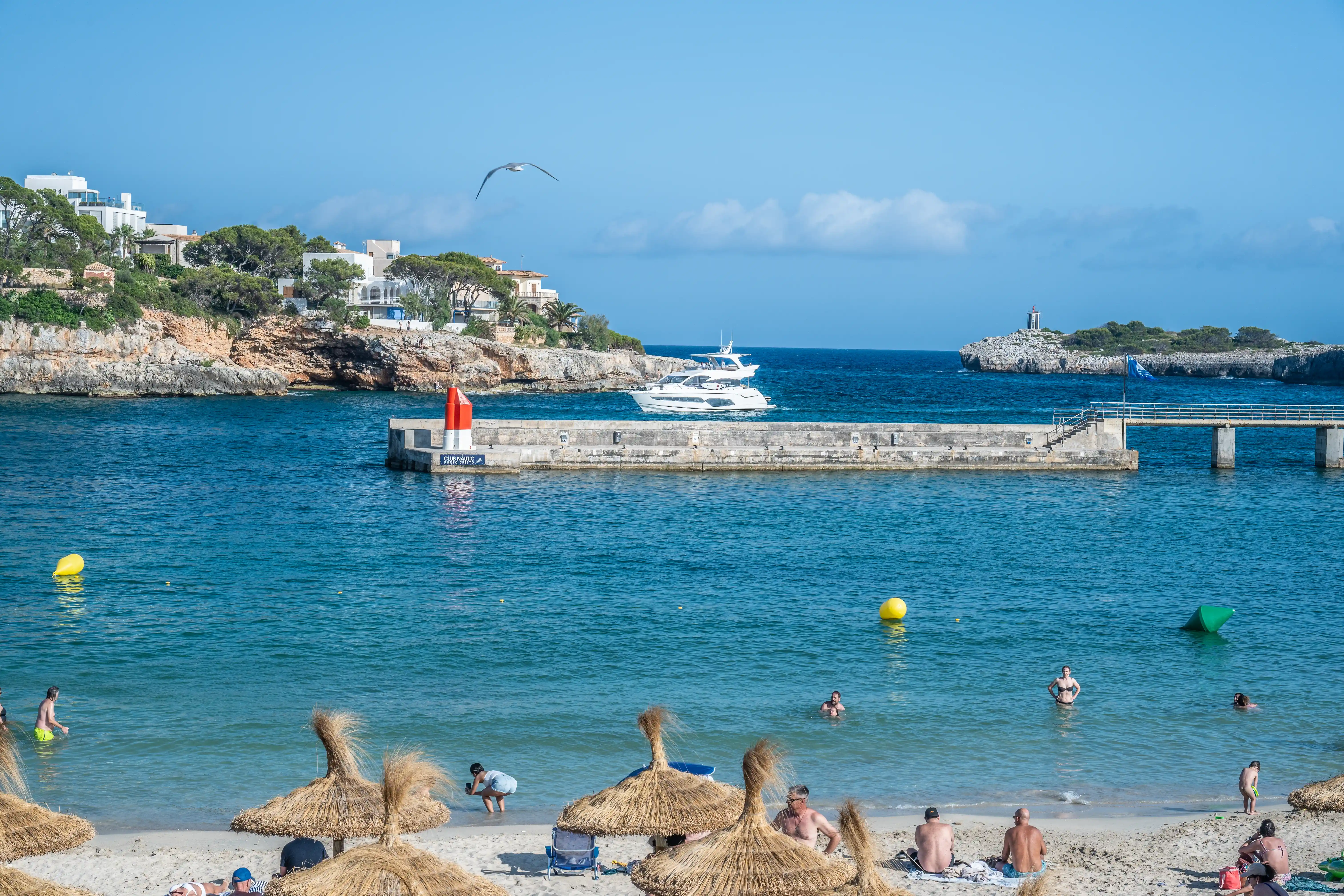 A scenic view of a bustling beach with people swimming and relaxing, a white yacht sailing in the blue sea, and coastal buildings on a cliffside under a clear blue sky.