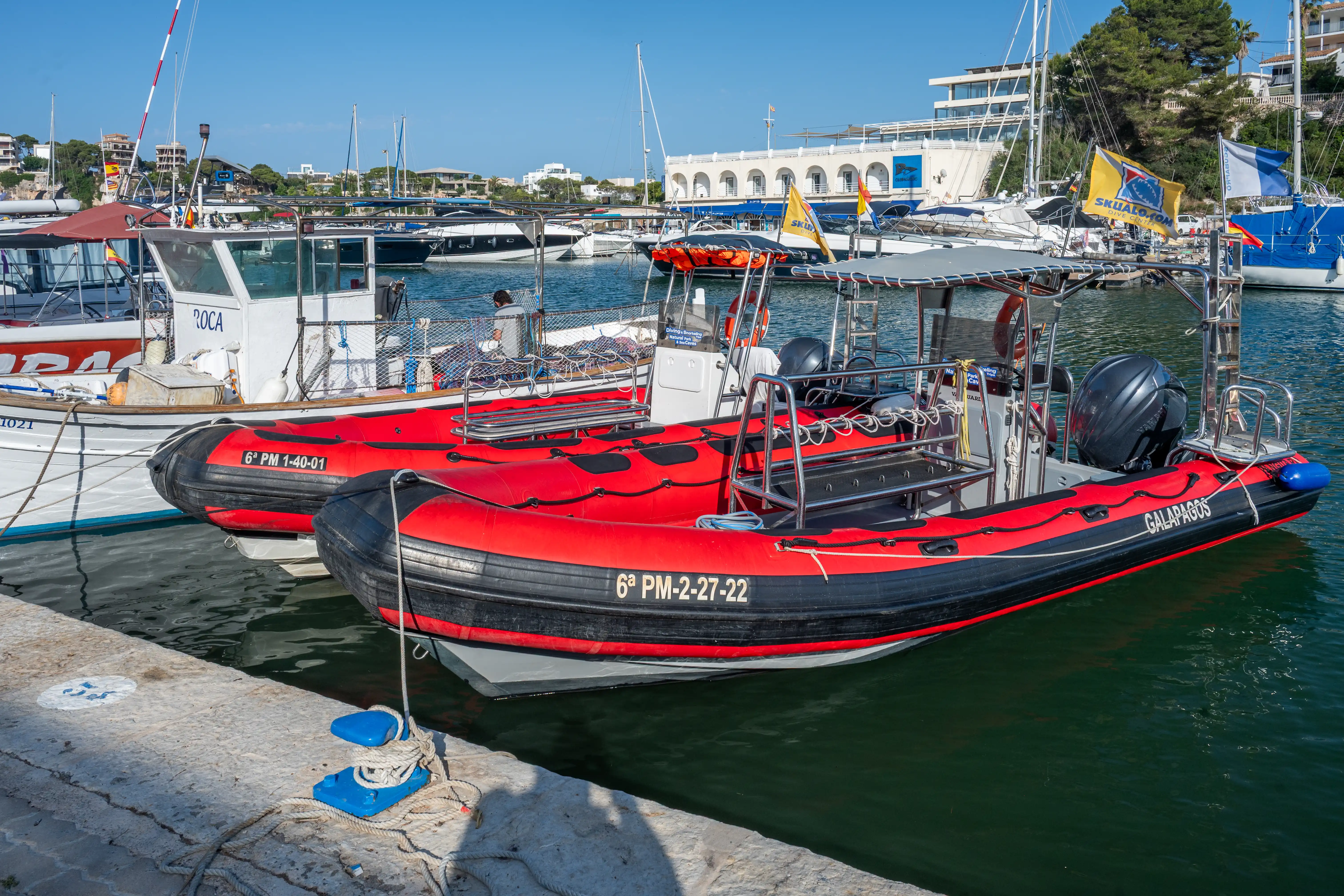 Two bright red rigid inflatable boats, one named GALAPAGOS, are moored in a harbour with other vessels and buildings in the background under a sunny blue sky.