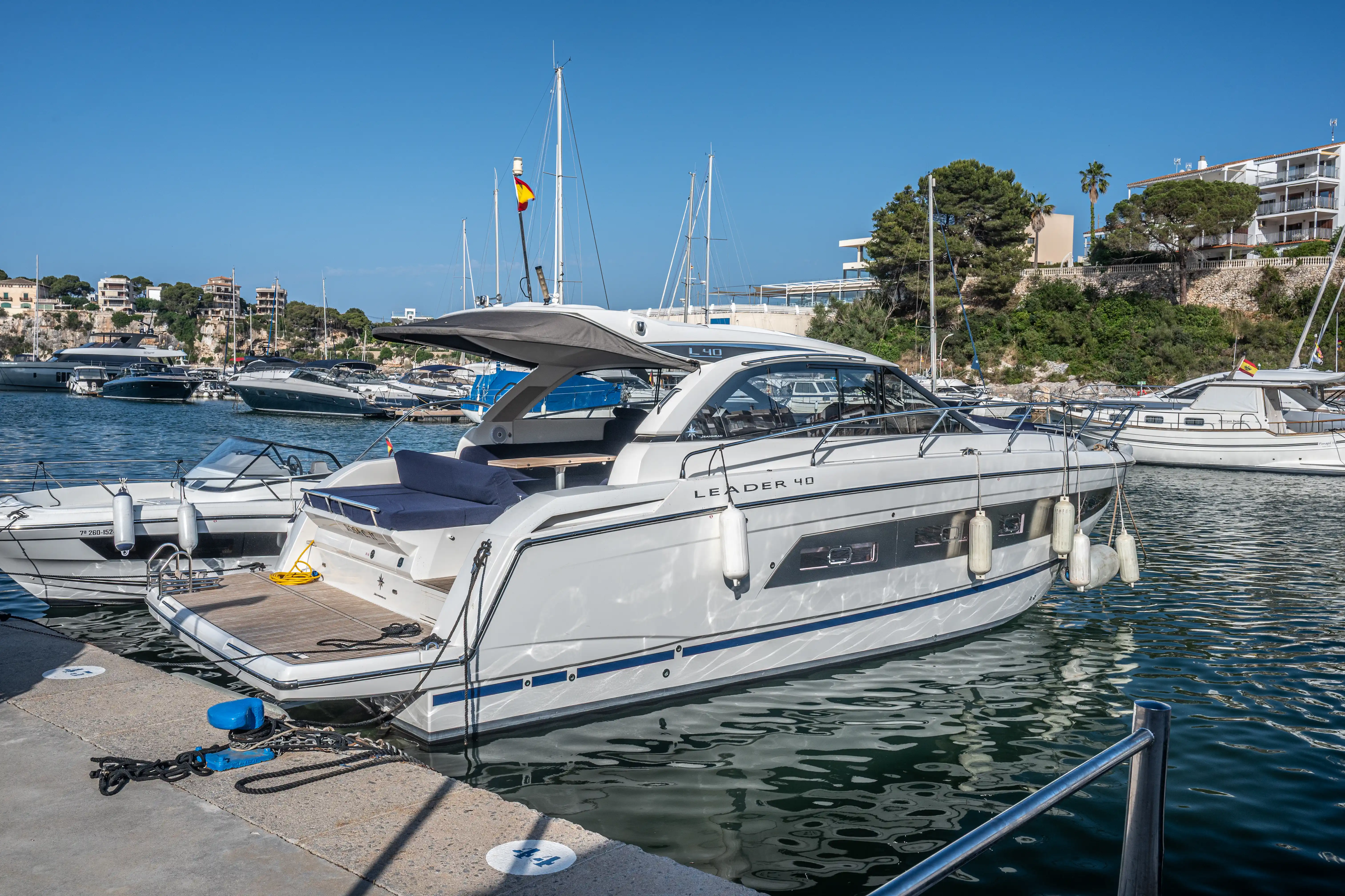 White motor yacht named LEADER 40 docked in a marina with other boats and buildings on a hill in the background under a clear blue sky.