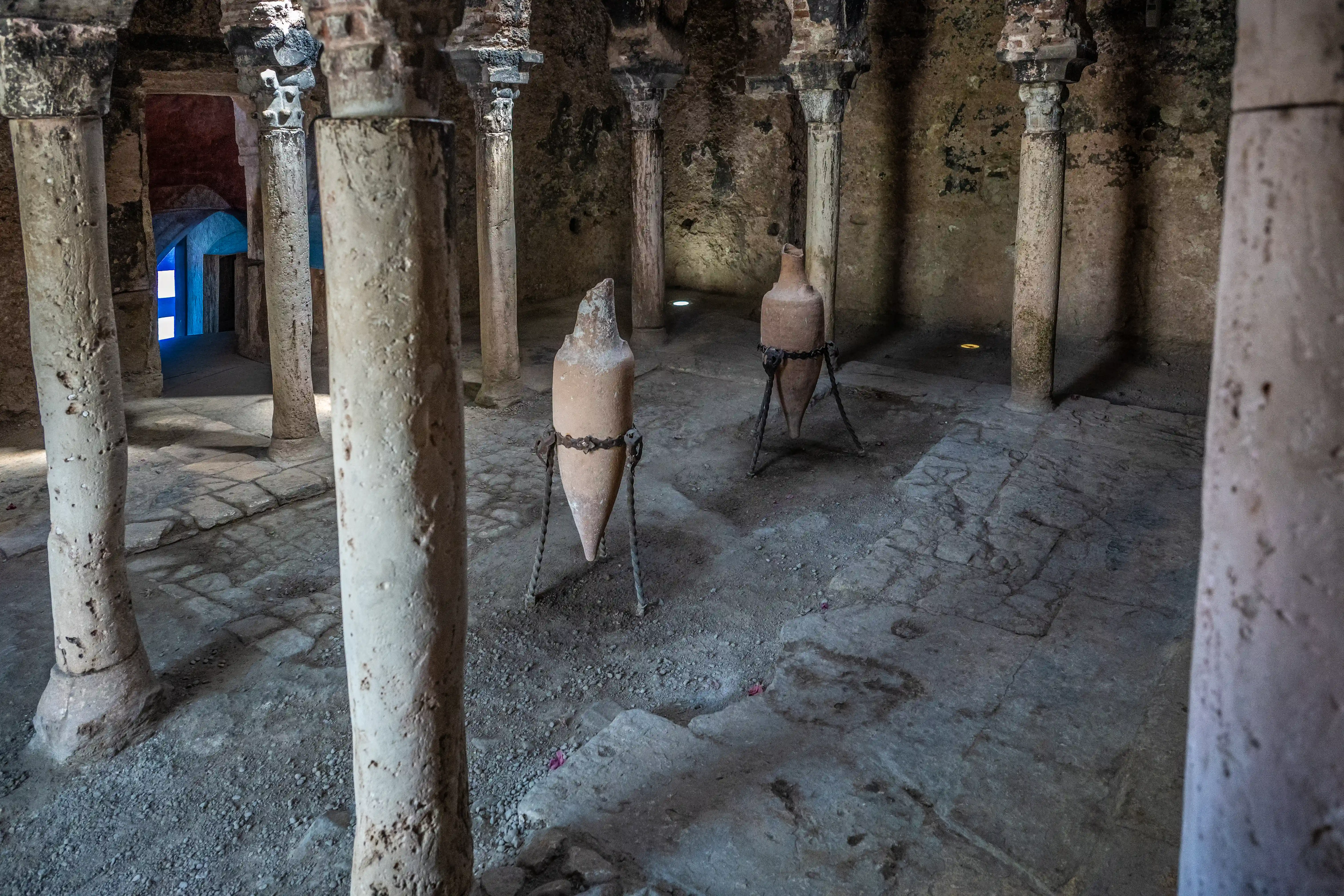 Interior view of an ancient stone chamber with several large, pointed terracotta amphorae displayed on metal stands. The room is supported by thick stone columns, and the floor is made of stone slabs. In the background, an archway is visible with bright blue light emanating from it.
