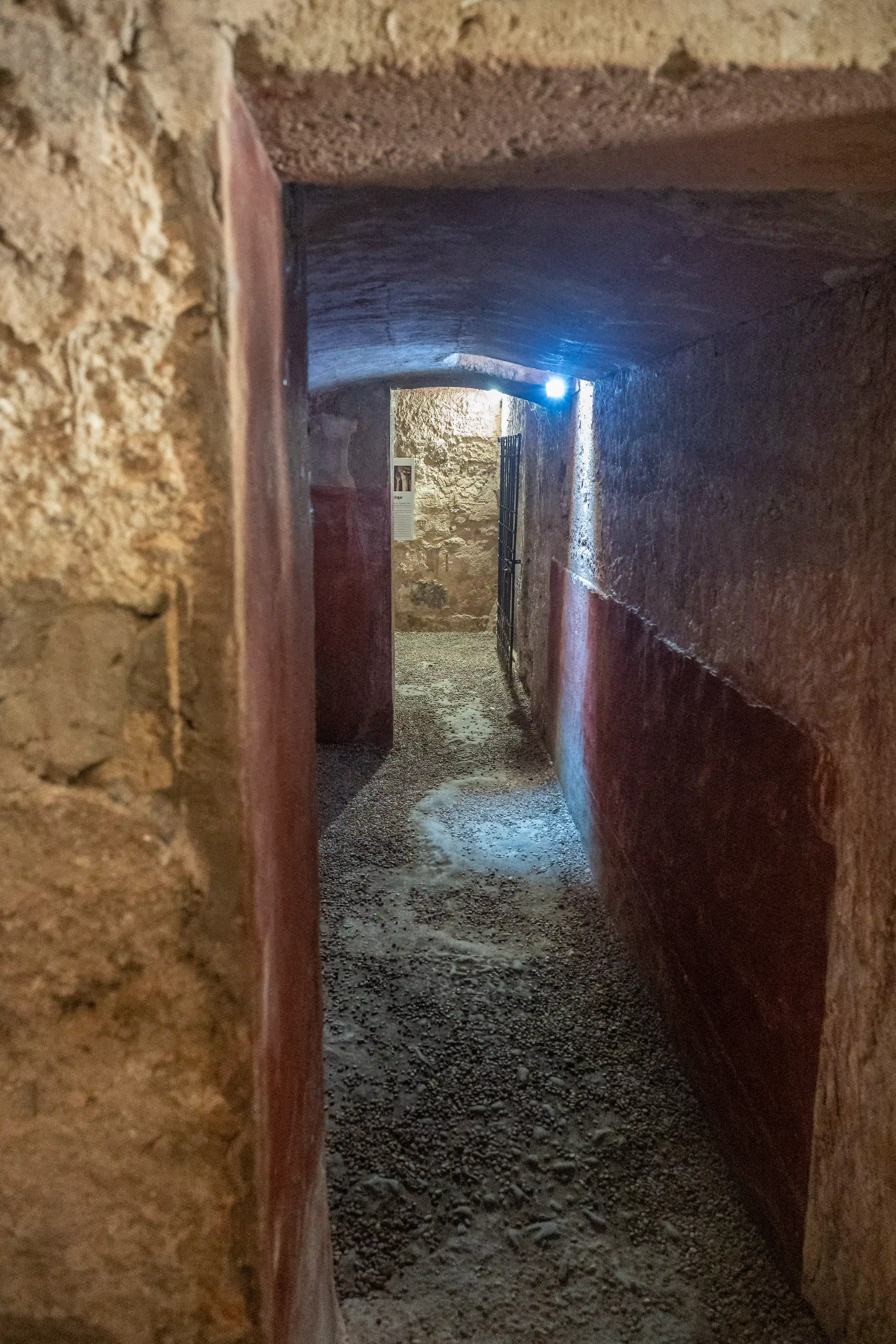 A narrow, dimly lit corridor constructed from rough stone walls. The corridor features a red painted band along the lower portion of the walls and a gravel floor. A metal gate is visible at the far end, beyond which lies another stone-walled area with a framed informational display. Artificial lighting illuminates the passage.