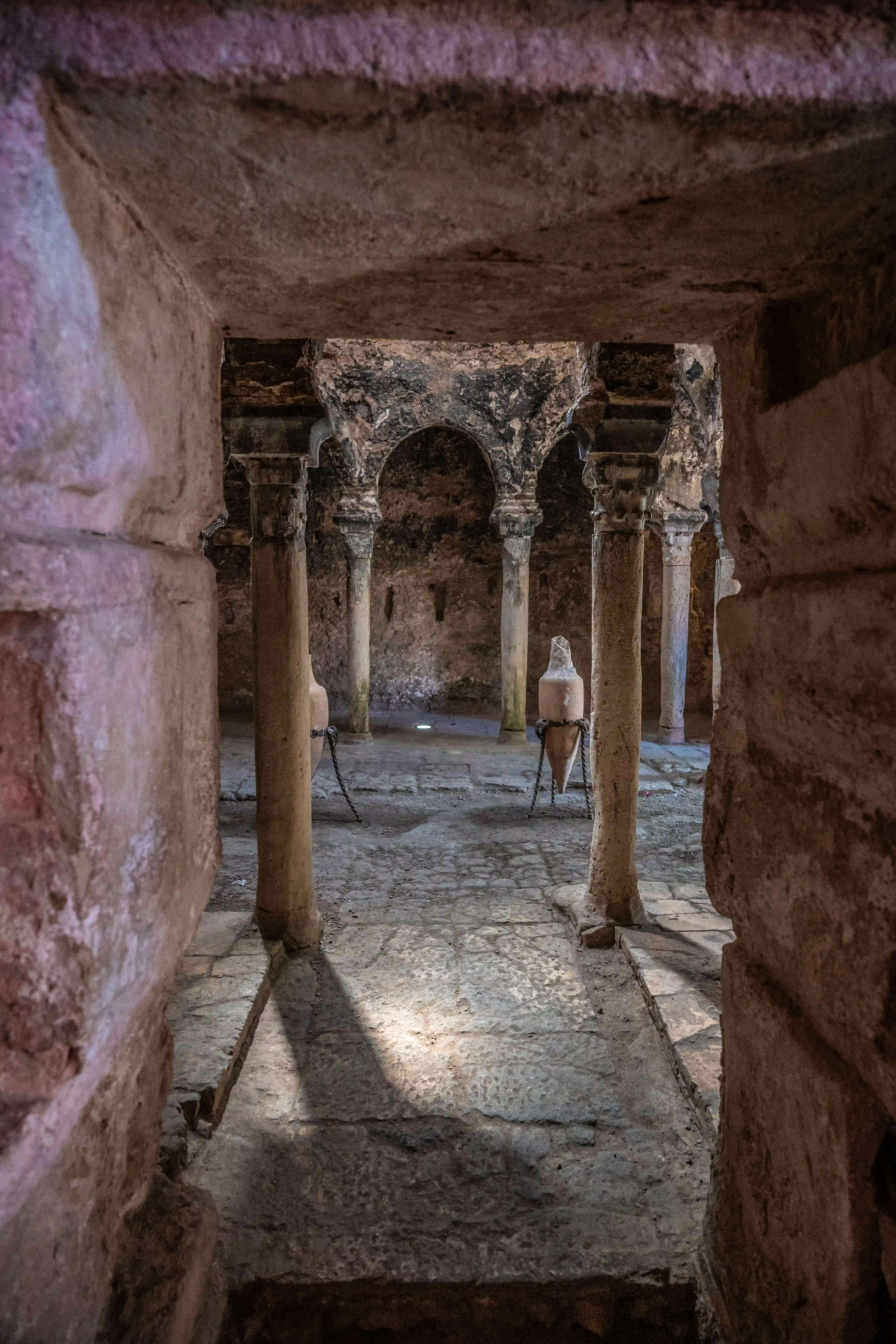 A view through a stone archway into an ancient chamber with stone columns and two large terracotta amphorae on display.