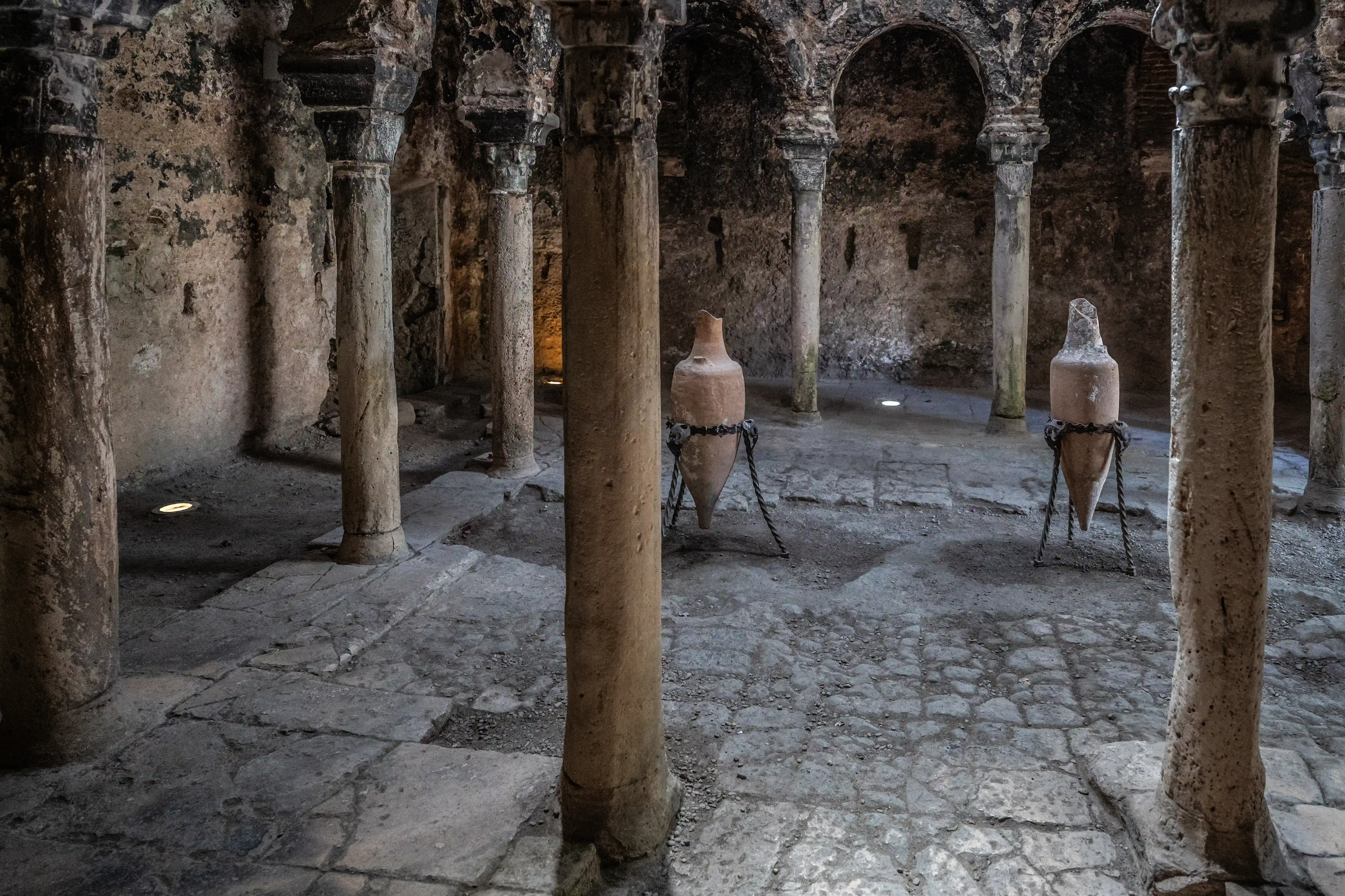 Interior view of a stone chamber with large, weathered columns and arched ceilings, featuring two ancient terracotta amphorae displayed on stands secured by chains.