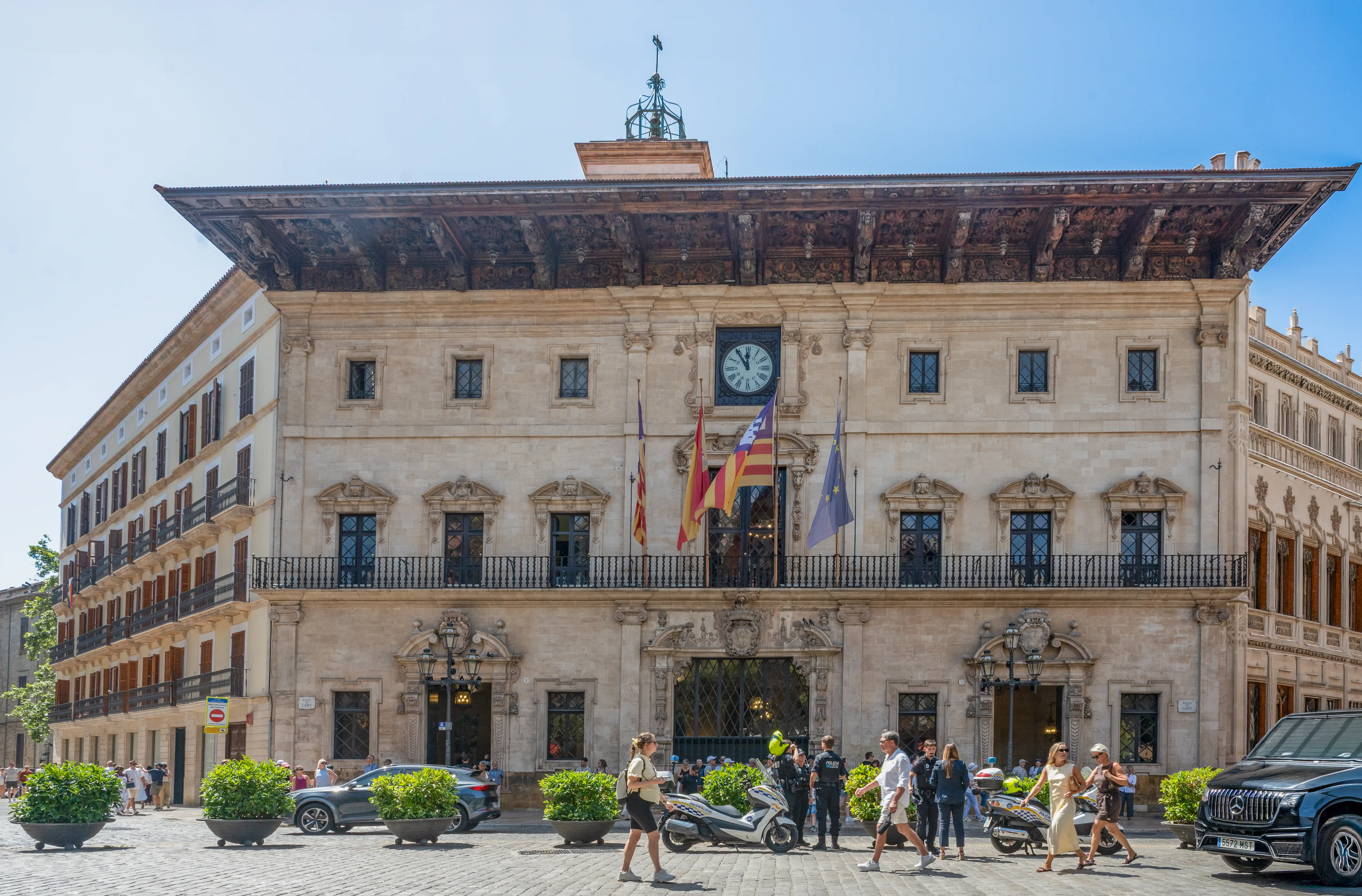 A grand, light-coloured stone building with a large clock and flags, viewed from a cobblestone plaza with people, vehicles, and greenery on a sunny day.