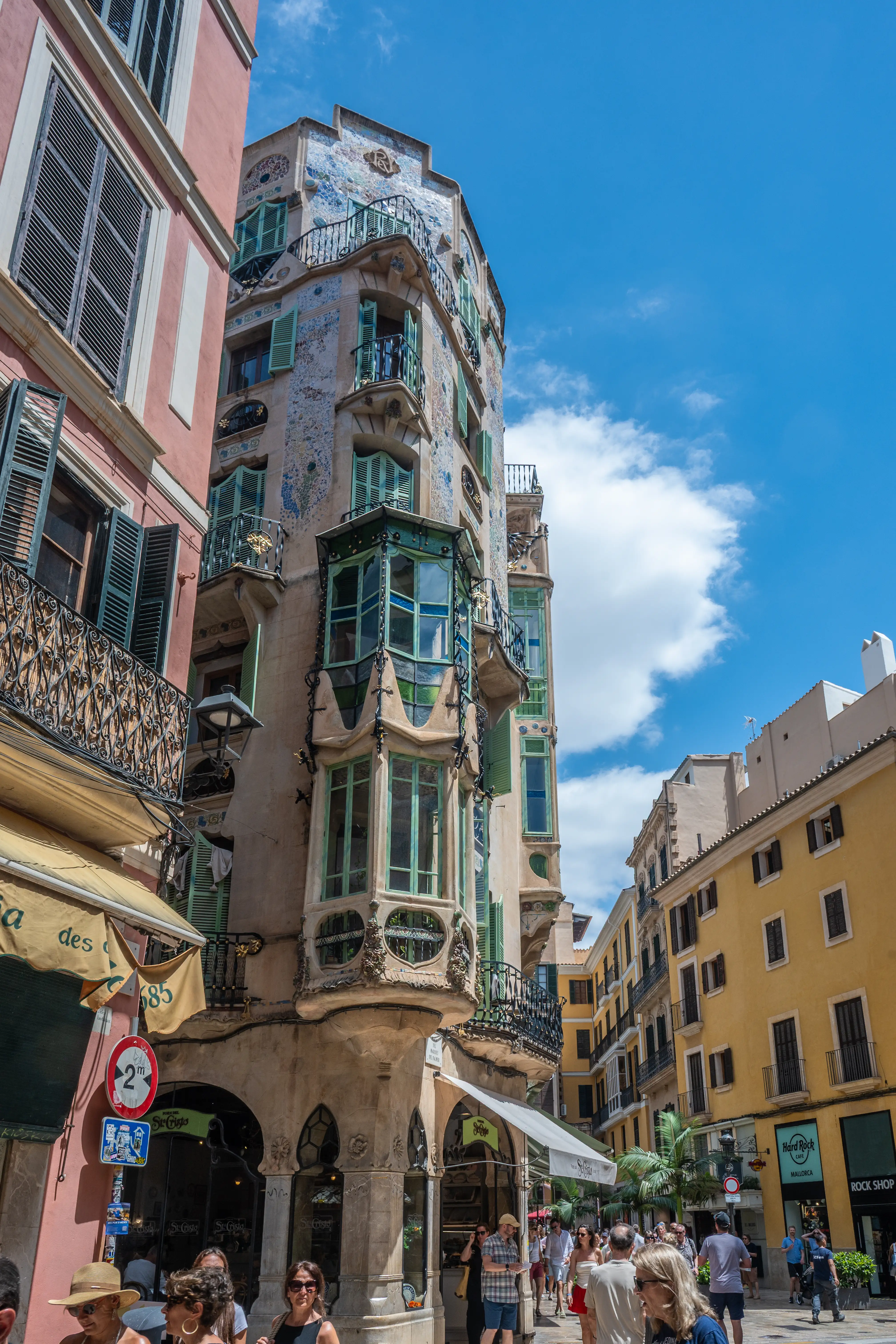  A narrow street in a European city with an ornate, multi-story building featuring mosaic tiles and green bay windows. People are walking by shops, with a bright yellow building on the right.