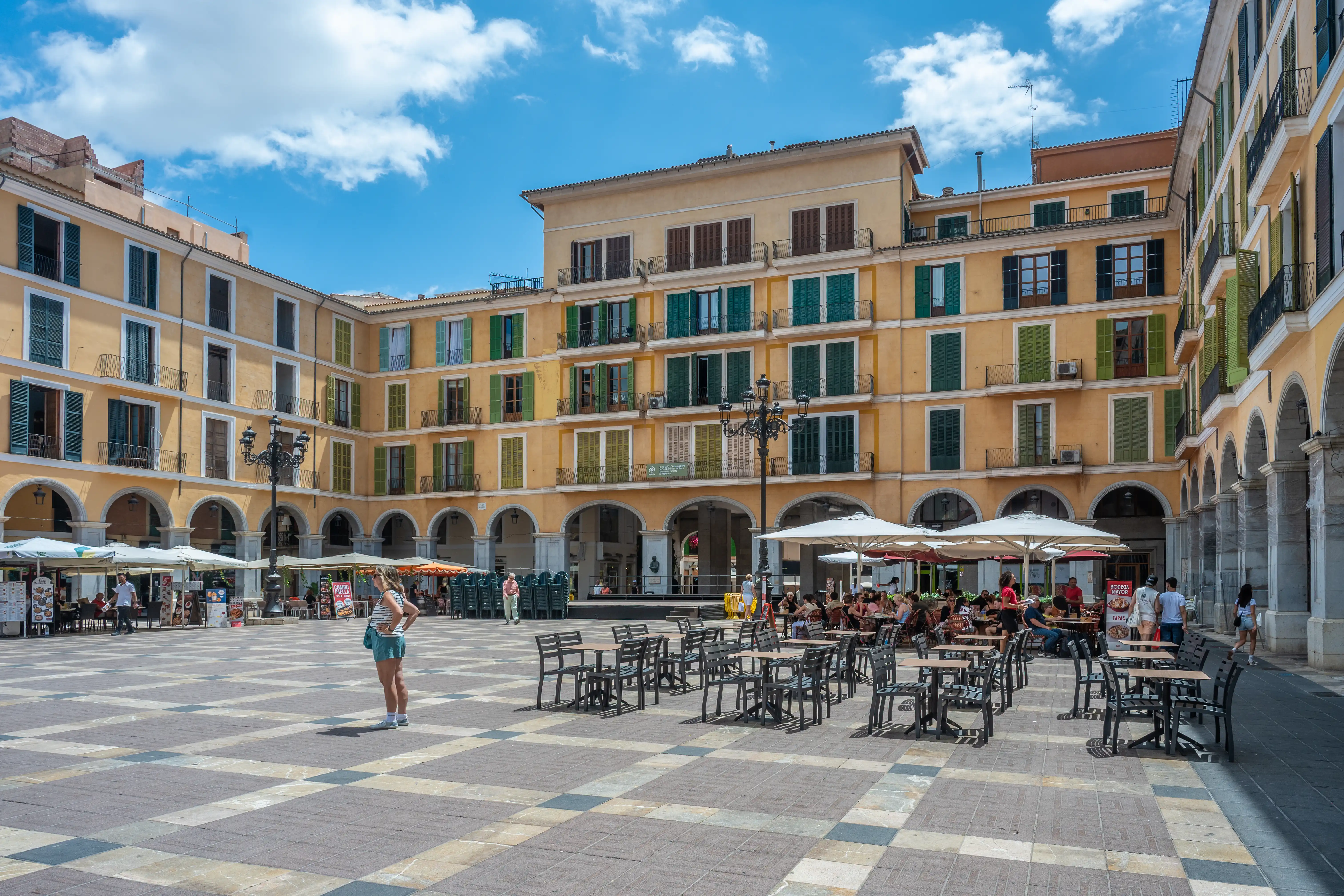 A sunny, wide plaza paved with checkered tiles, surrounded by bright yellow buildings with arcades on the ground floor and windows with green shutters on the upper floors. Outdoor seating areas for cafes are visible, with people seated and walking around. A lone woman stands in the foreground, facing away from the camera.