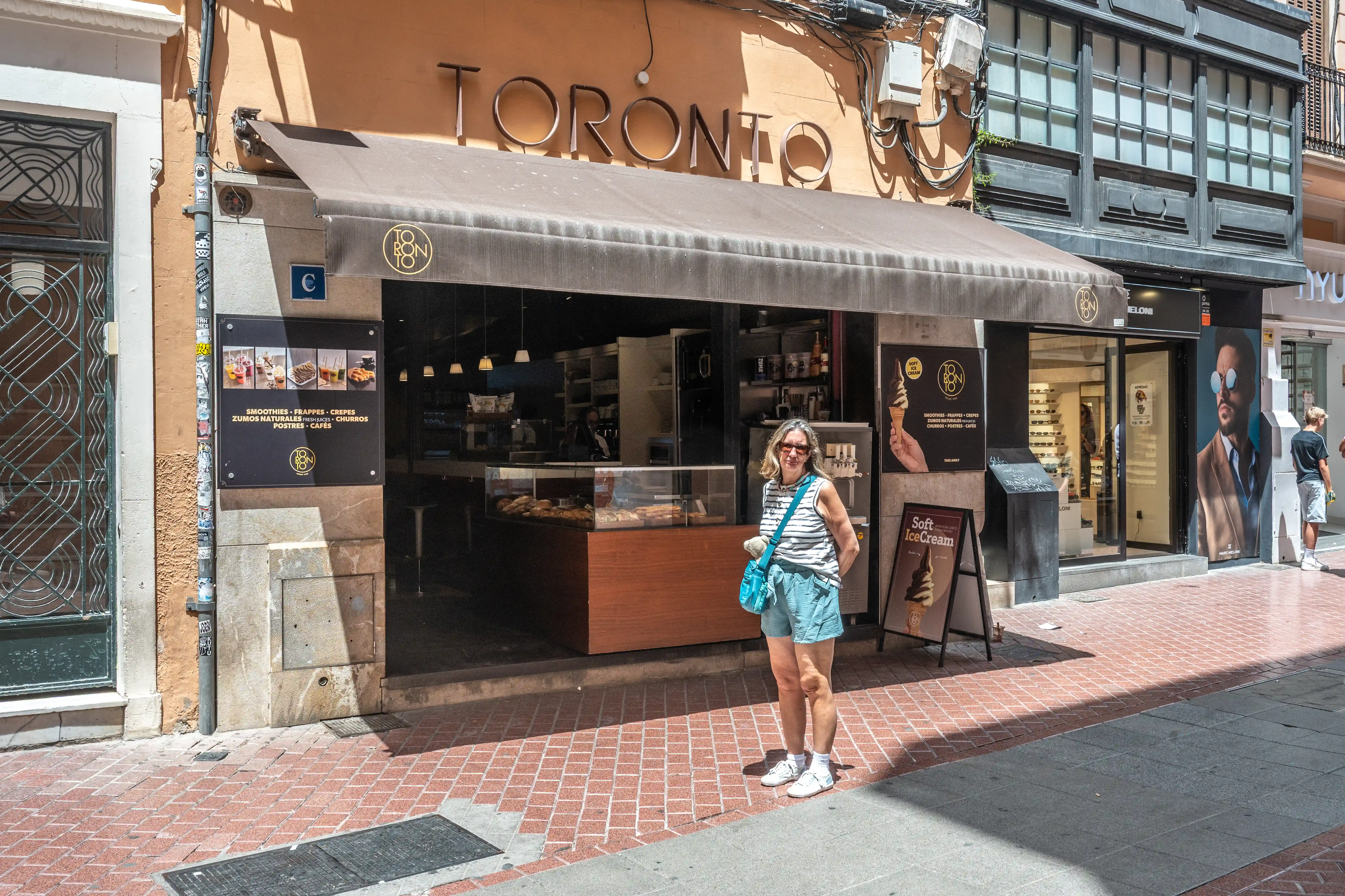 A woman with blonde hair, wearing a striped t-shirt and blue shorts, smiles at the camera in front of a cafe with a large sign reading 