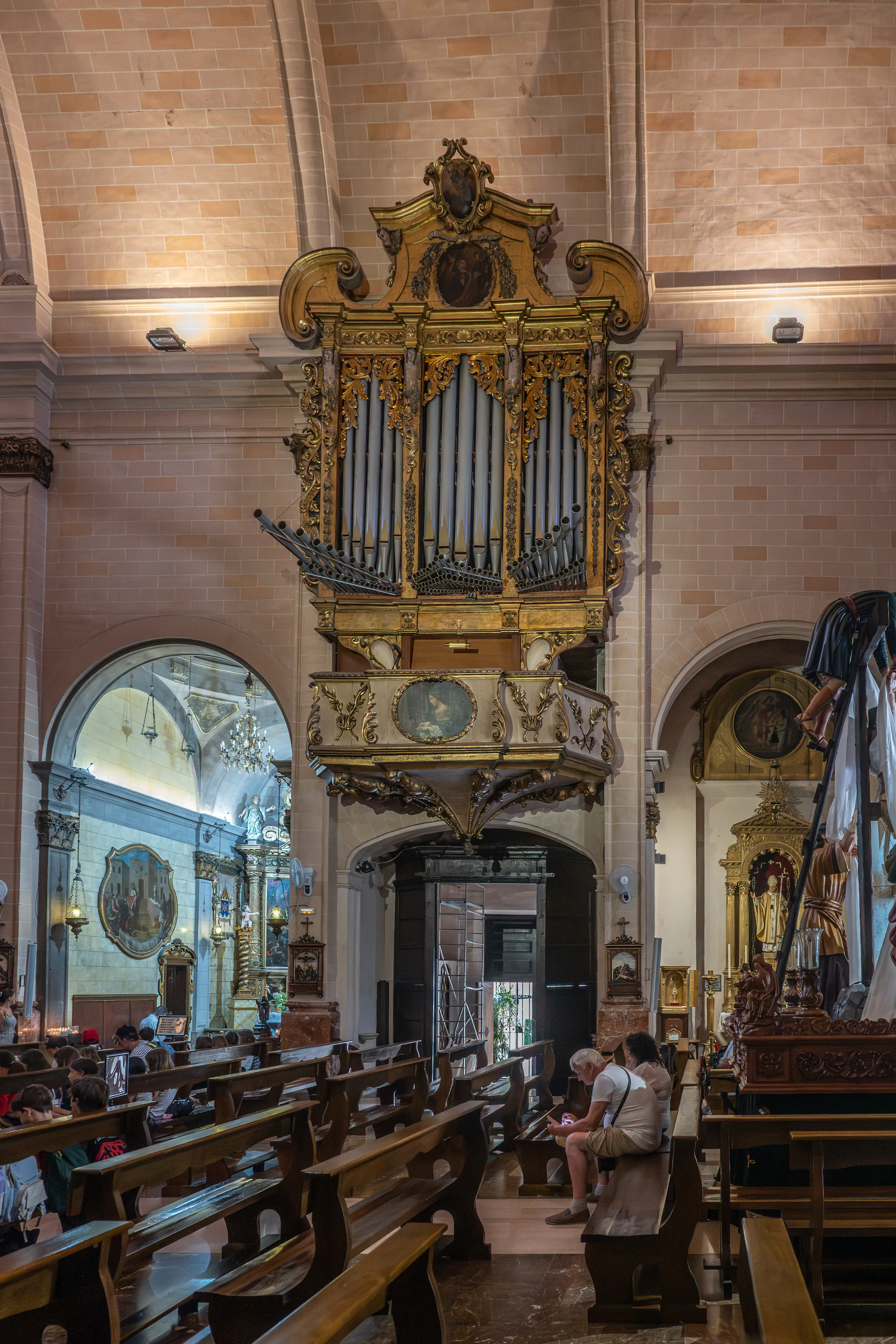 Interior view of a church nave with rows of wooden pews, a large, highly decorated golden pipe organ on the wall, and a religious tableau on the right side.