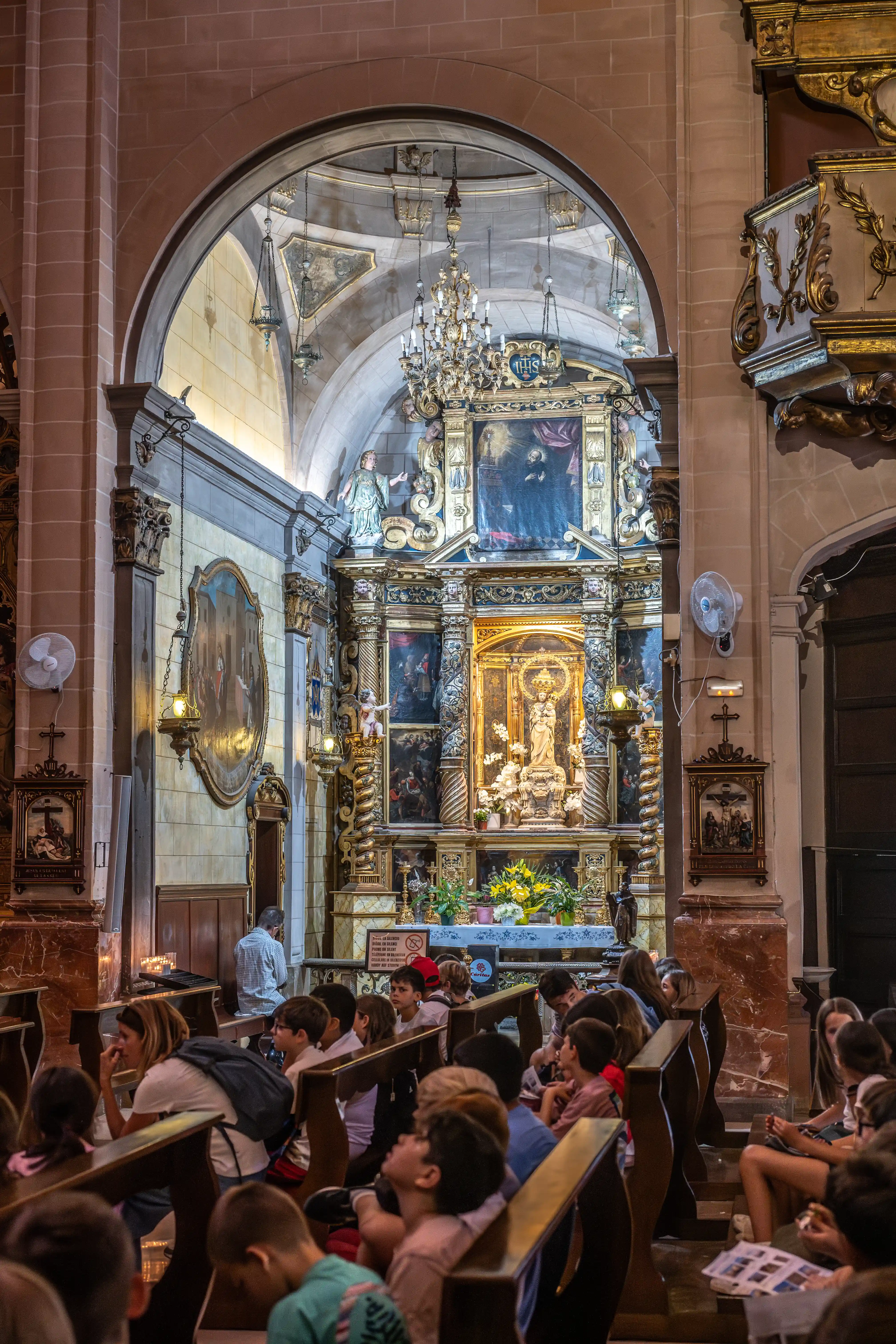 A wide shot of a church interior, showing a highly decorated gold altar with a statue of Mary and Jesus at its centre. In the foreground, a group of people, including many children, are seated in wooden pews, facing the altar.