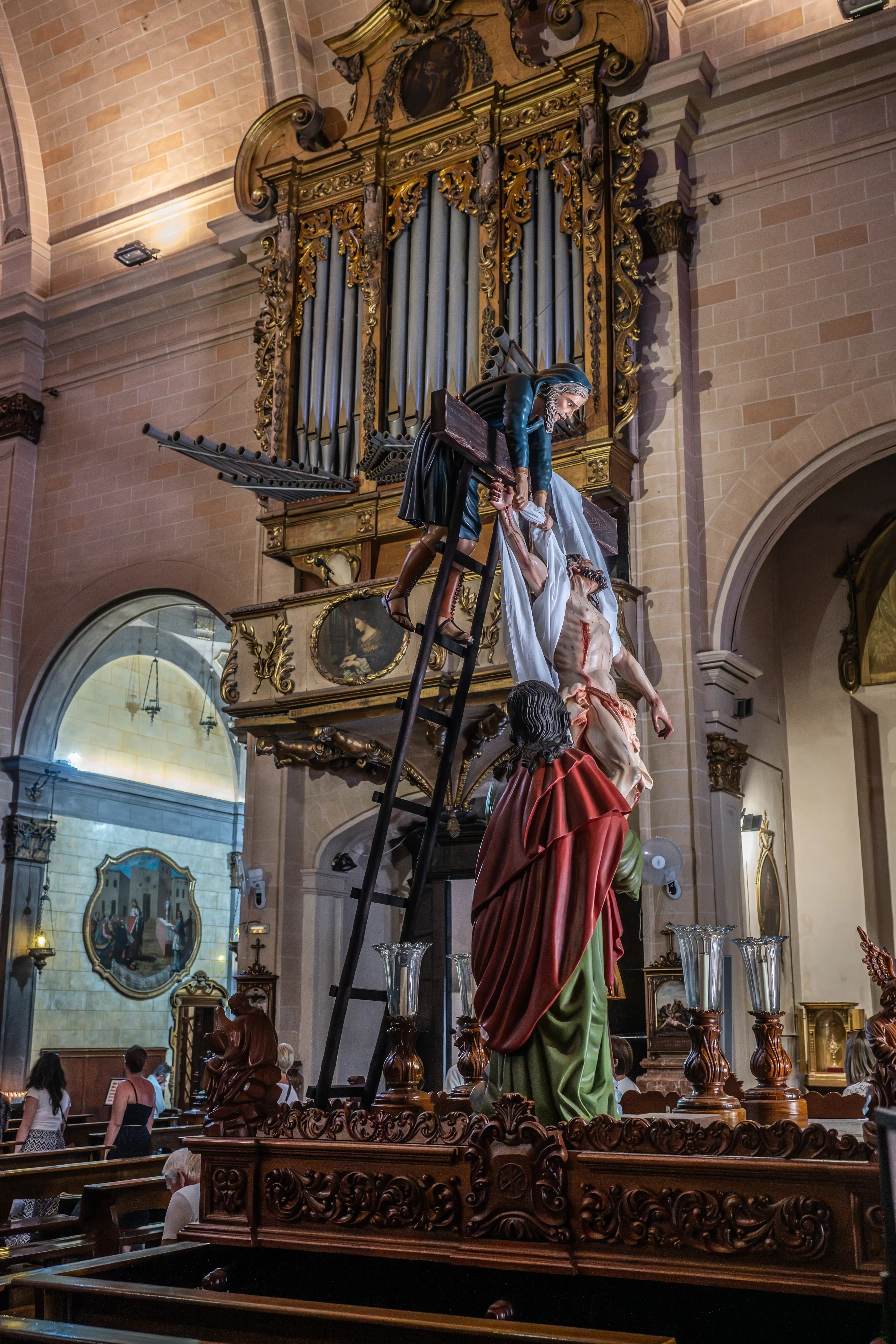 A religious statue depicting the lowering of Jesus from the cross inside a church, with an ornate organ in the background and people sitting in pews.