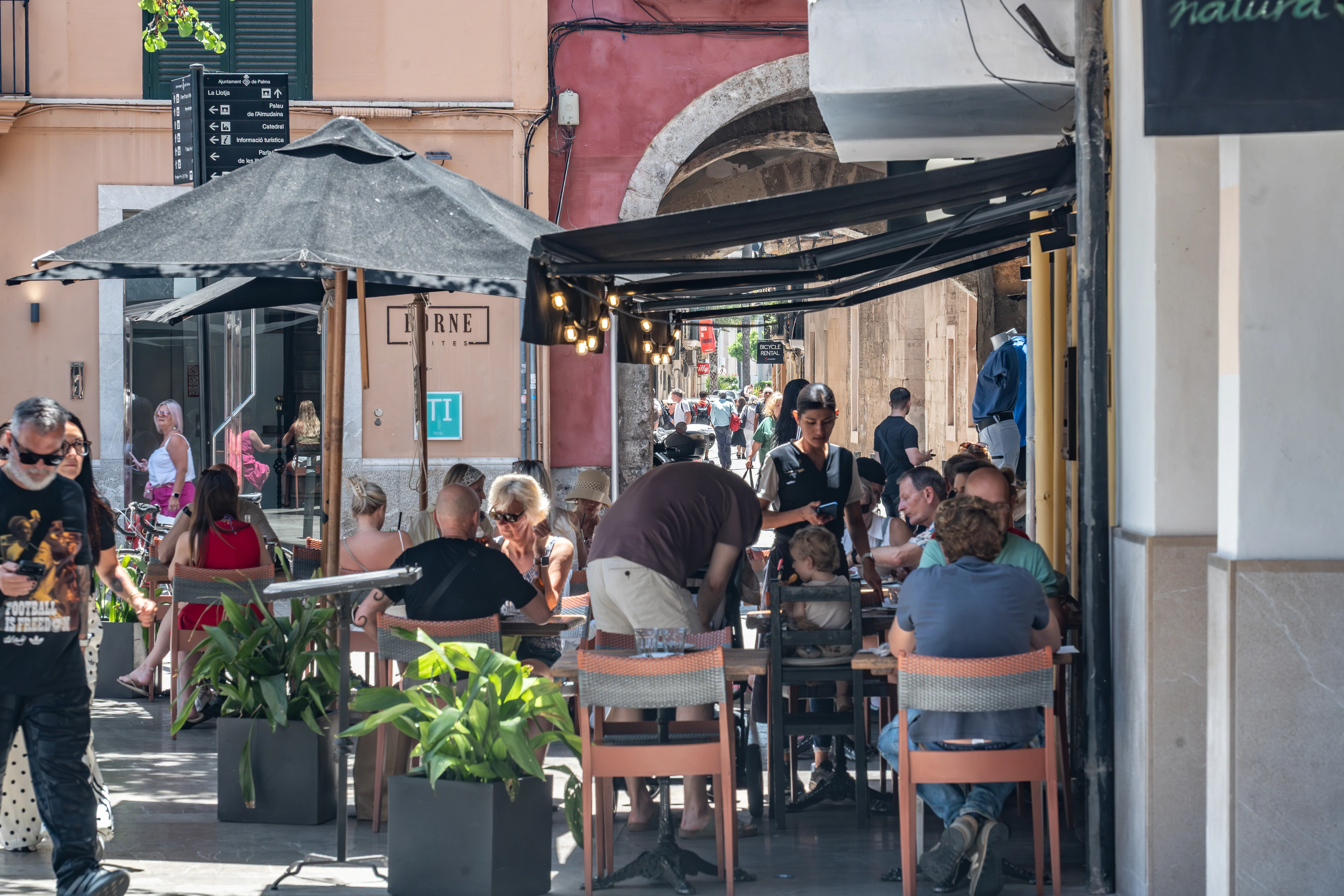 A group of people are seated at tables outside a cafe in Palma, Spain. Umbrellas and awnings provide shade. A waiter is serving customers, and there are plants in pots. The buildings in the background are typical of a European city.