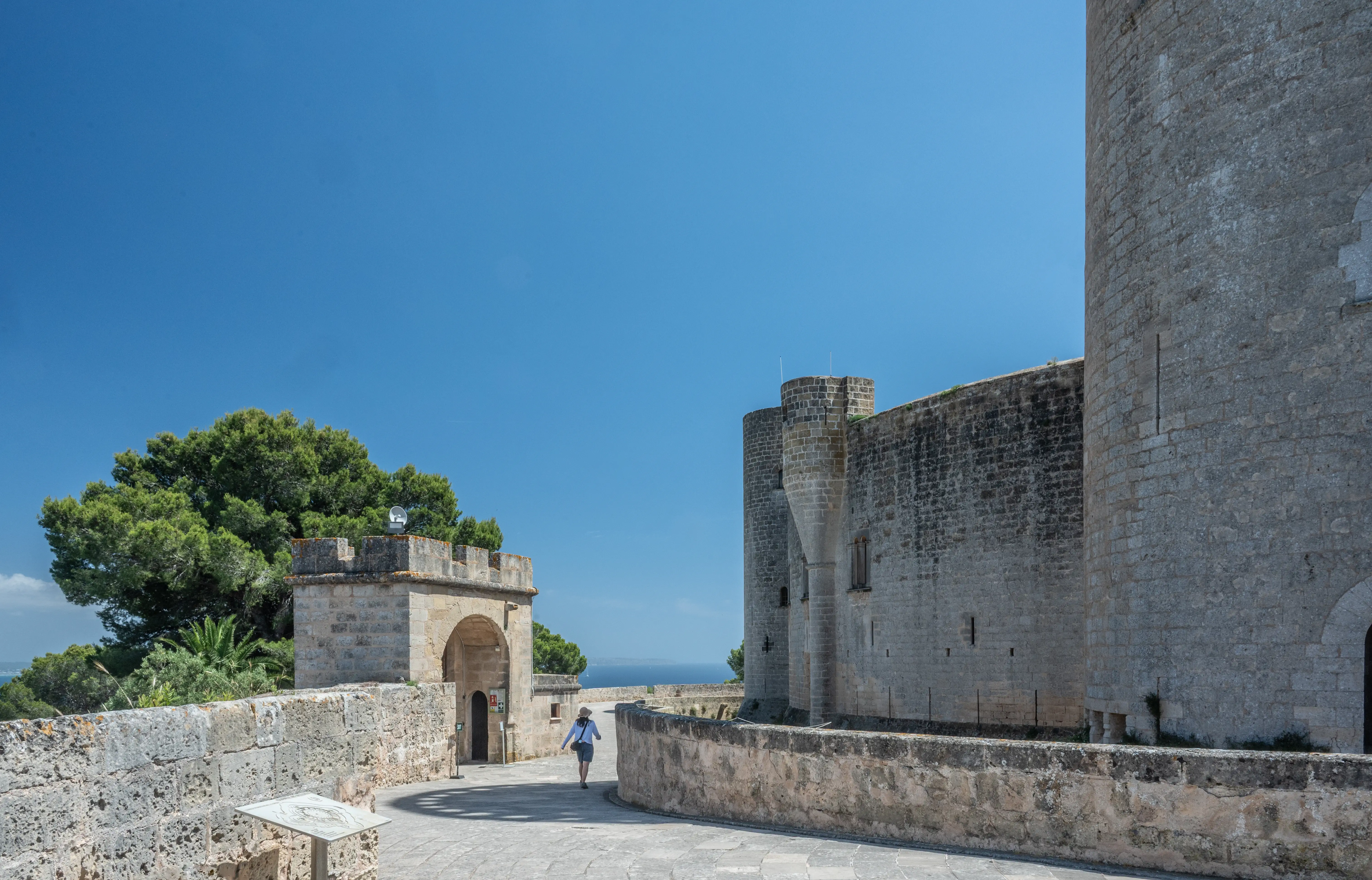 A person walks along a stone pathway leading towards an arched gateway in a medieval castle complex. Large stone towers and walls, a lush green tree, and a bright blue sky dominate the scene.