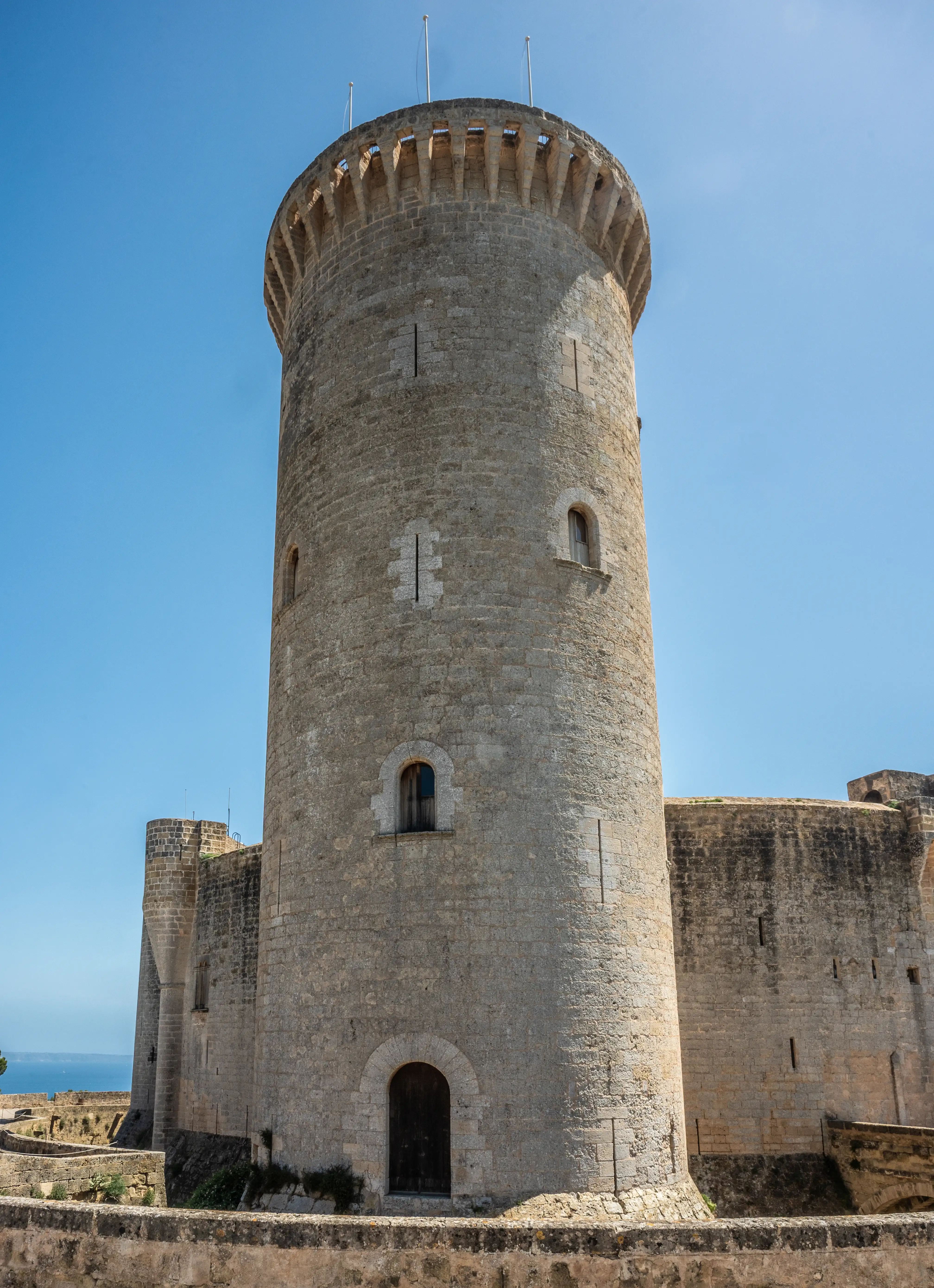 A towering ancient stone castle keep with narrow windows and battlements, set against a bright blue sky, with sections of castle walls and a glimpse of the sea in the background.