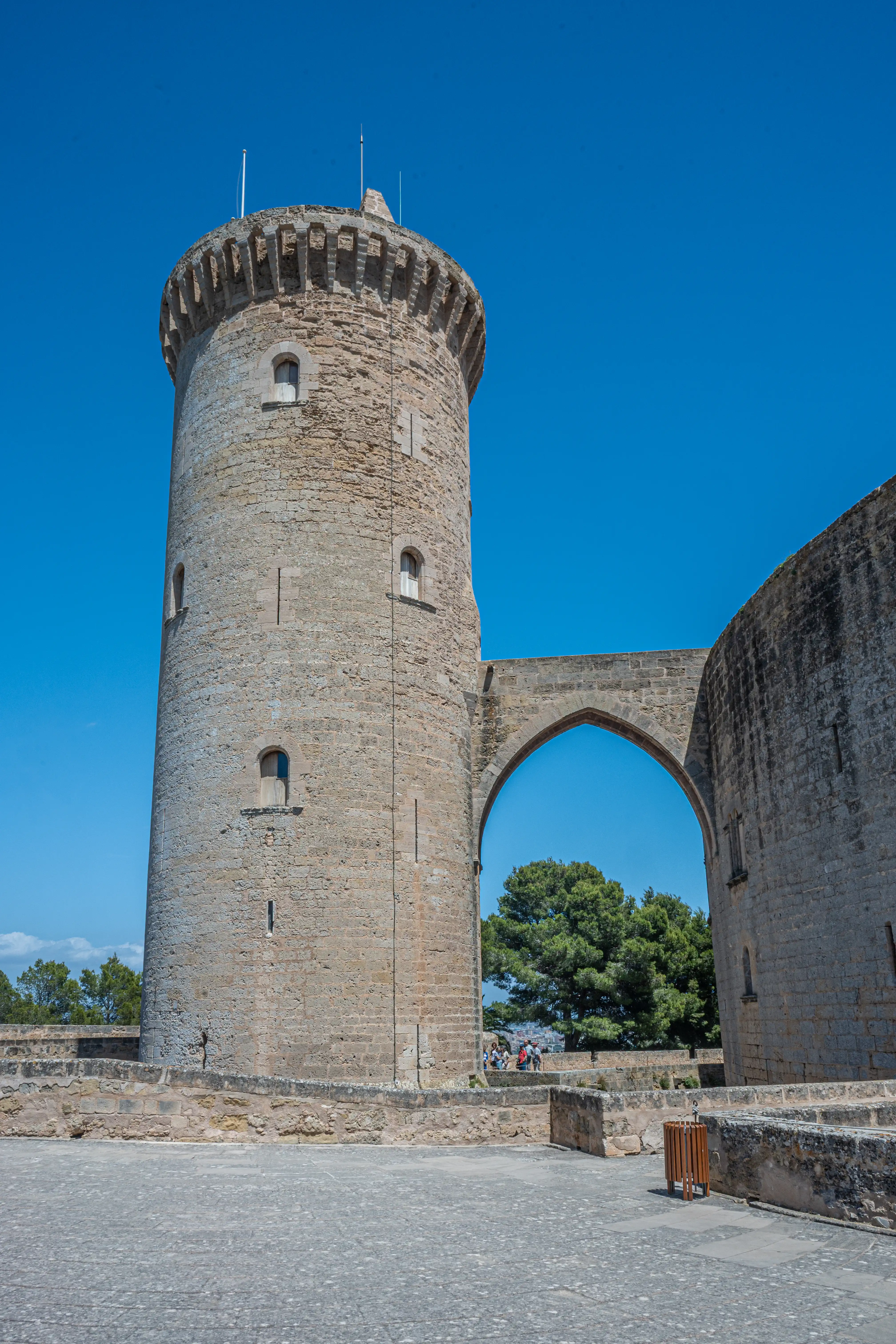 A circular stone tower connected by an arched bridge to a curved wall of a castle, under a clear blue sky.