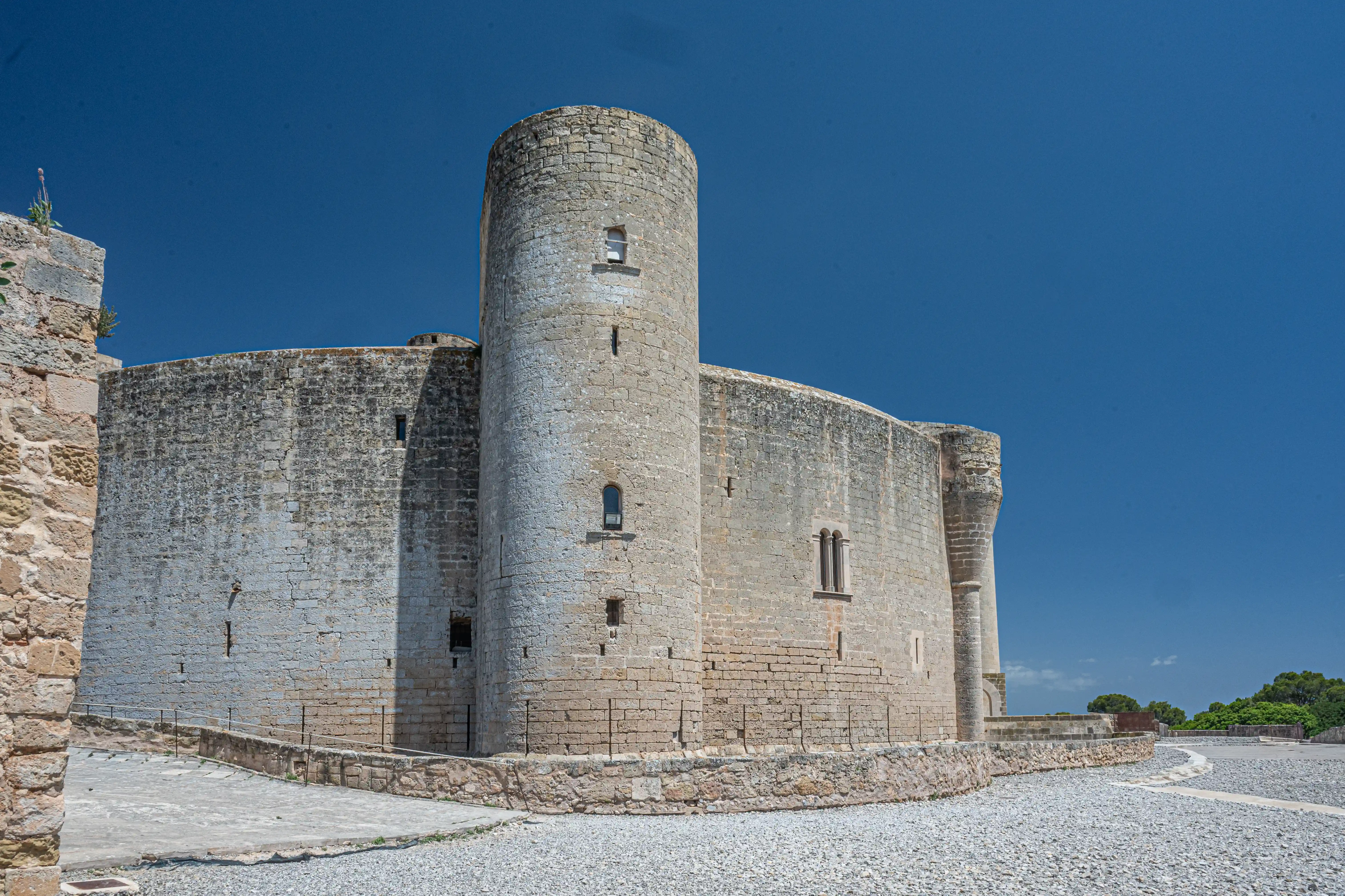 A large, circular stone castle with a tall tower and arched windows, set against a bright blue sky. The ground in front is covered in gravel and a paved path.