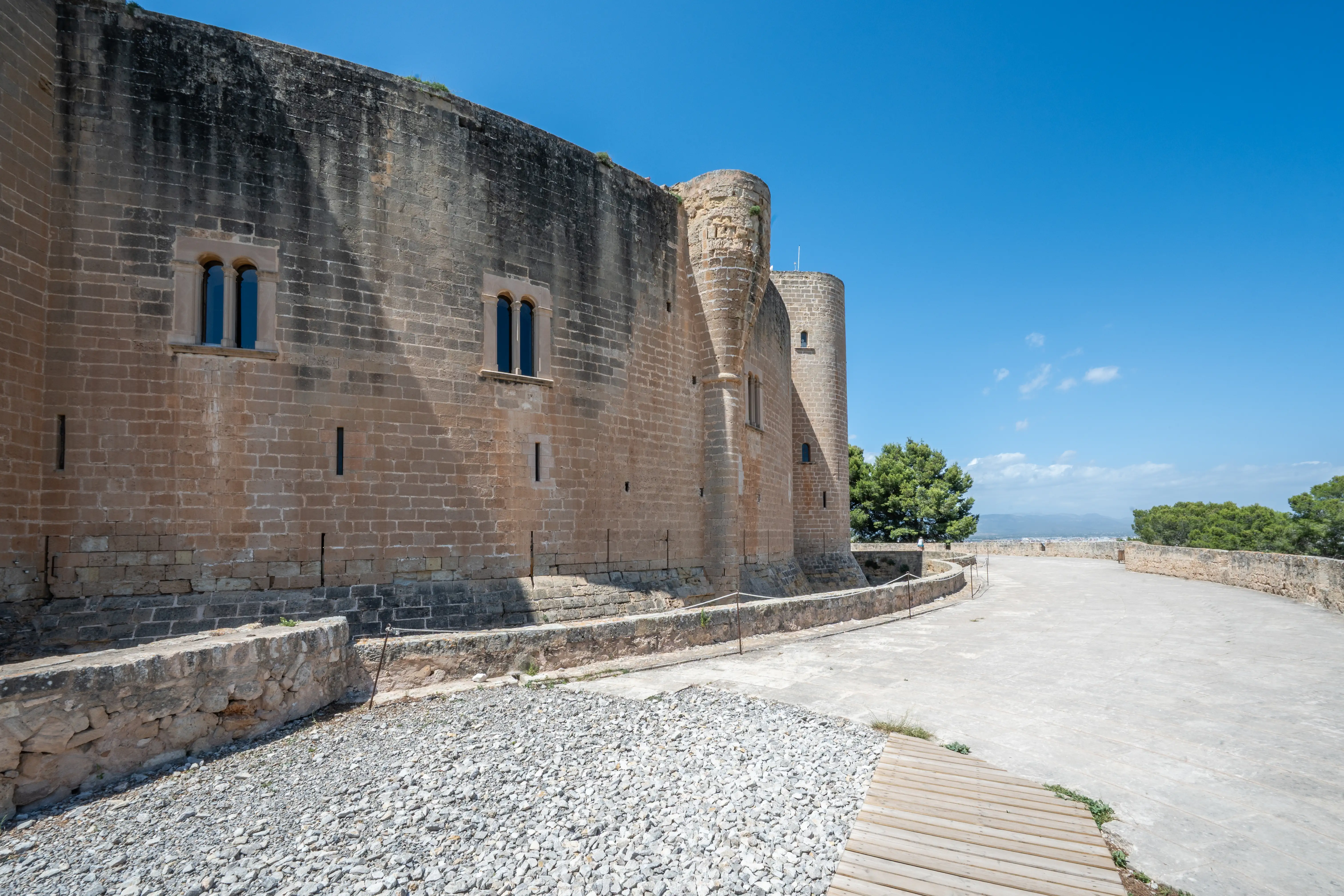 Wide shot of a weathered stone castle wall with arched windows and round turrets on a bright, sunny day, with gravel and wooden decking in the foreground and trees and distant landscape beyond.