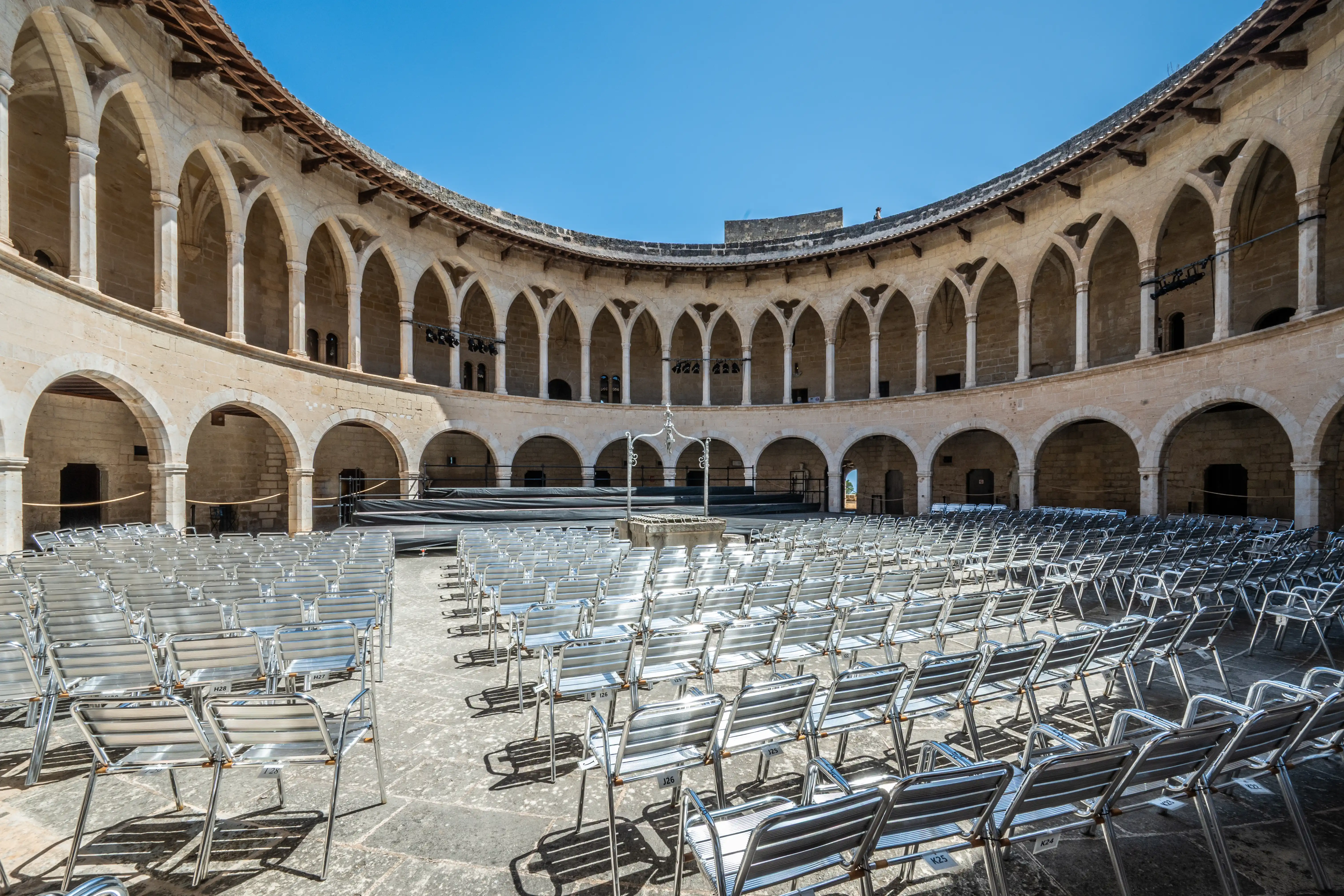 Rows of empty silver chairs are set up in the stone courtyard of a historic building with arched colonnades under a clear blue sky.
