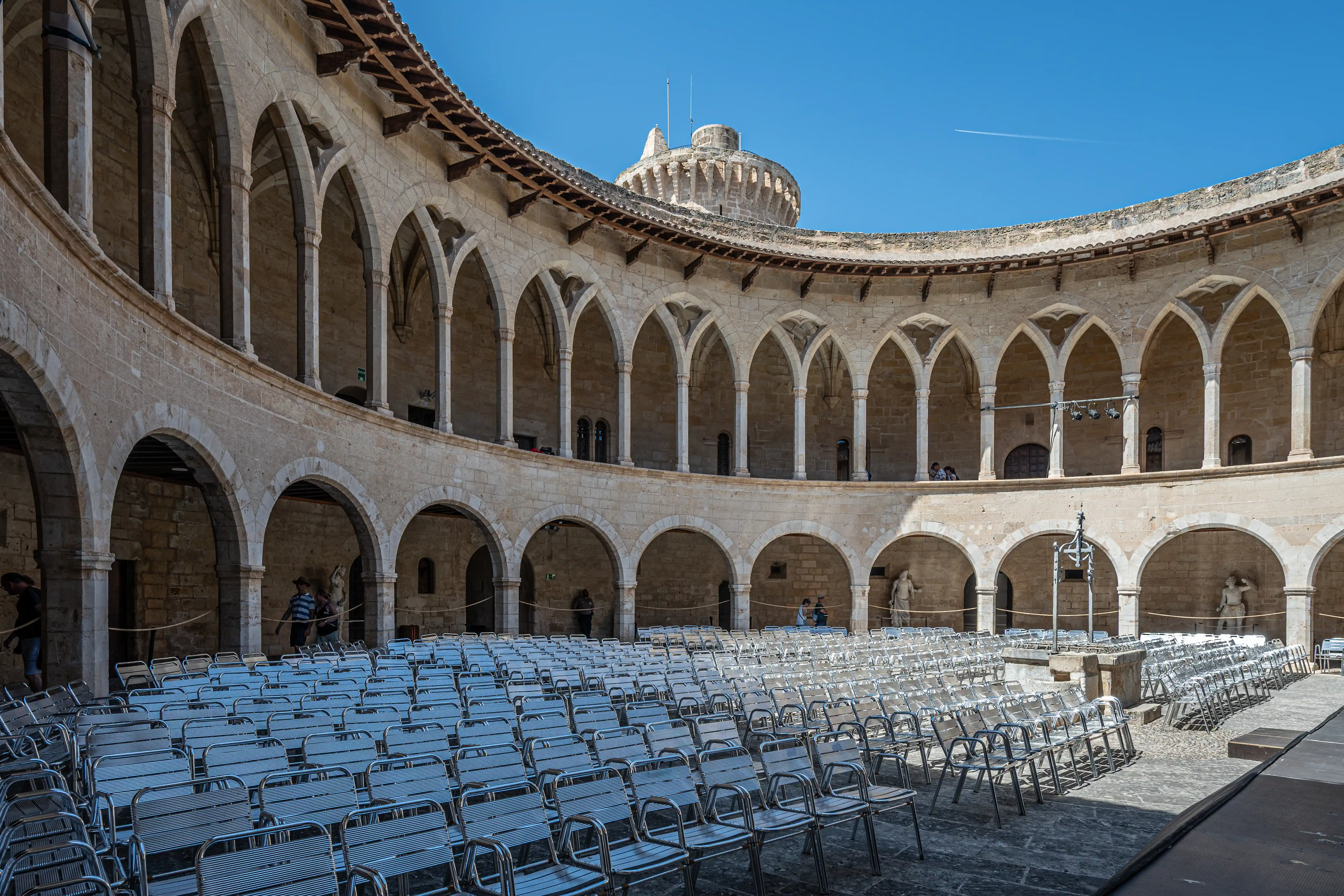 A wide shot of the courtyard of Castillo de Bellver, featuring numerous rows of empty silver chairs set up on paving stones. The palace's distinctive stone architecture, with numerous arched walkways and a circular tower, is visible behind the seating. A bright blue sky with a faint contrail from an aeroplane is overhead.