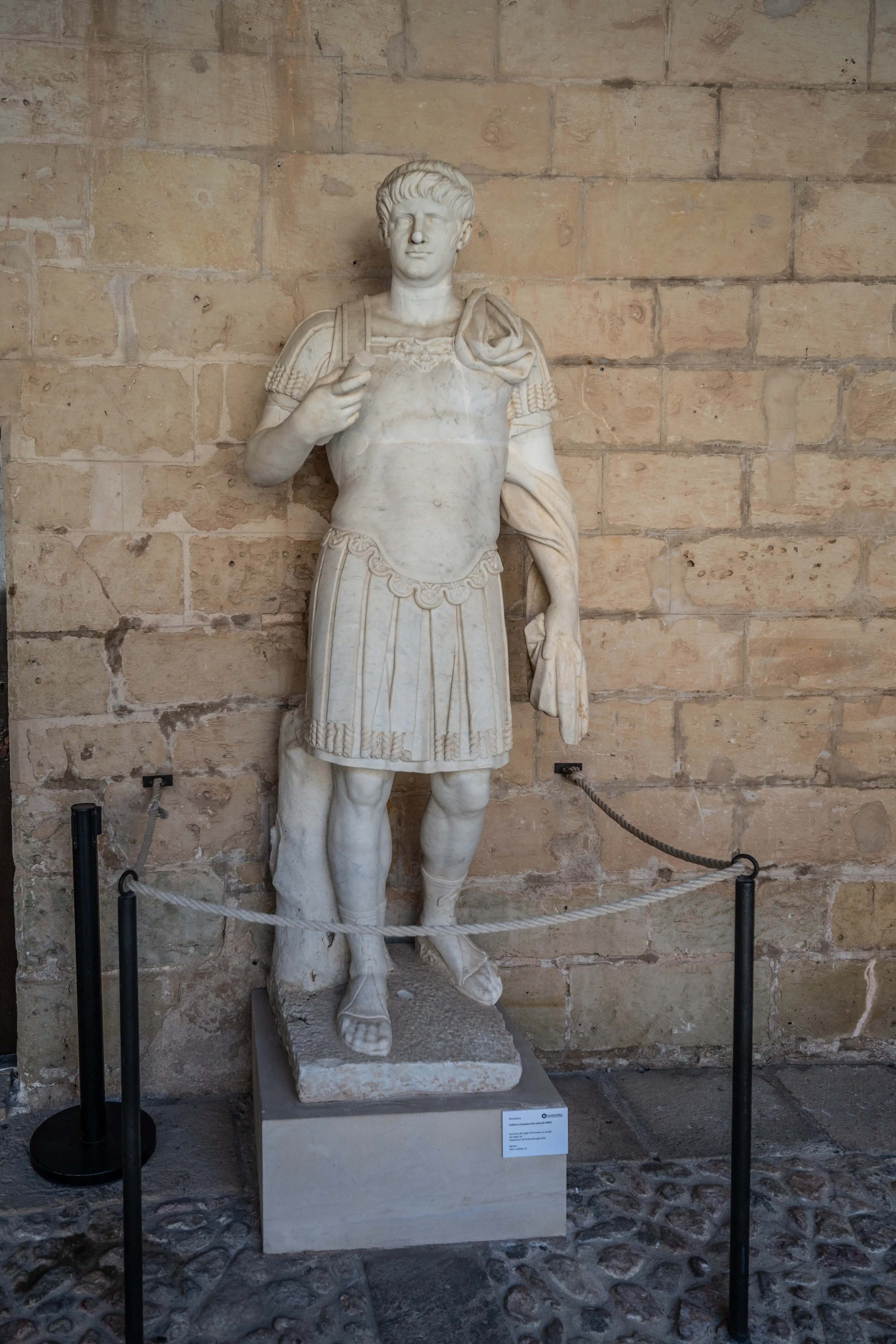 A white marble statue of a Roman emperor in armour, standing against a tan stone brick wall, with a rope barrier in the foreground.