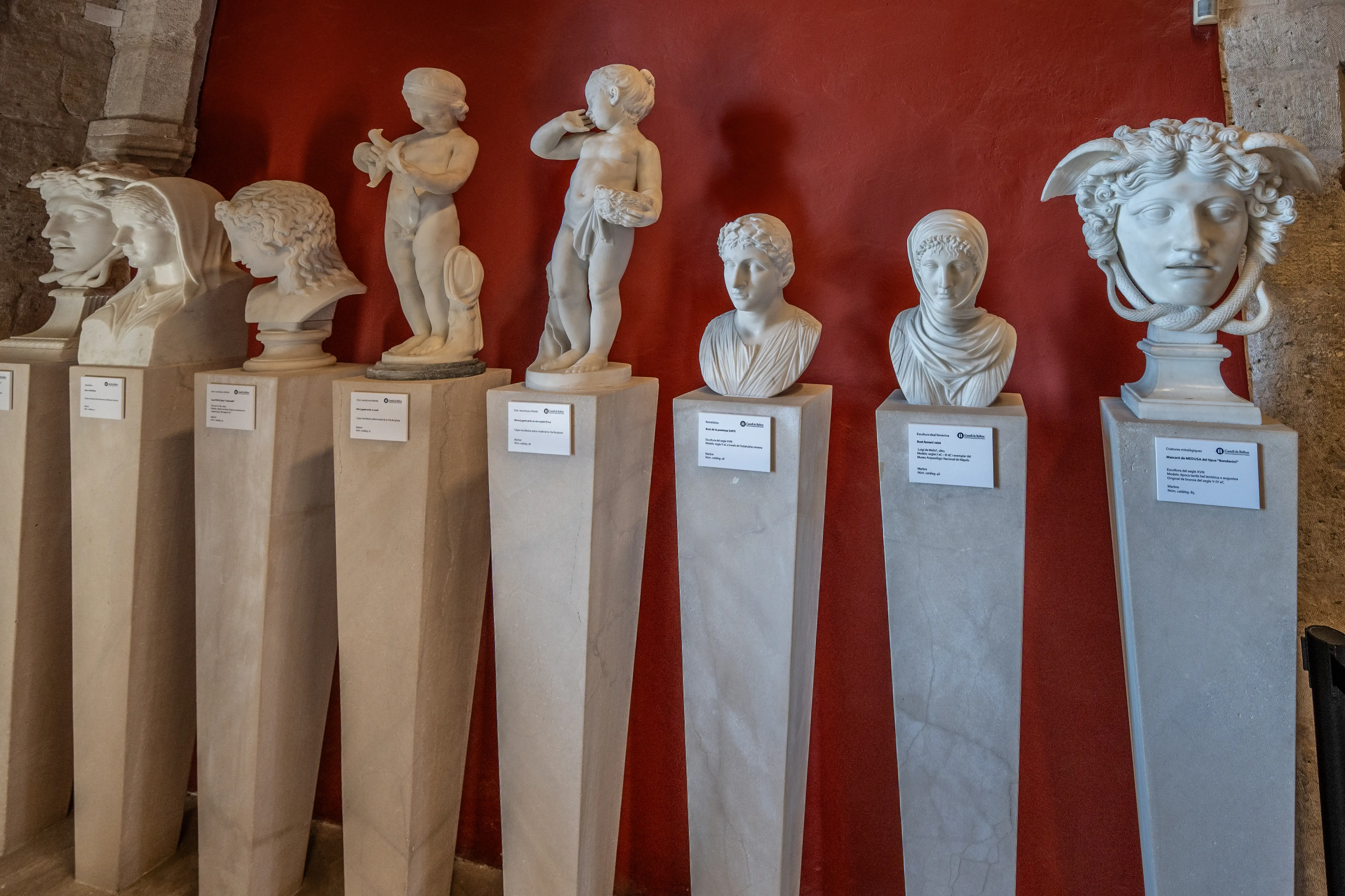 Several classical marble sculptures of heads and children are displayed on plinths against a vibrant red background. The sculptures vary in style, with some being detailed busts and others depicting cherubic figures. The lighting highlights the texture and form of the marble.