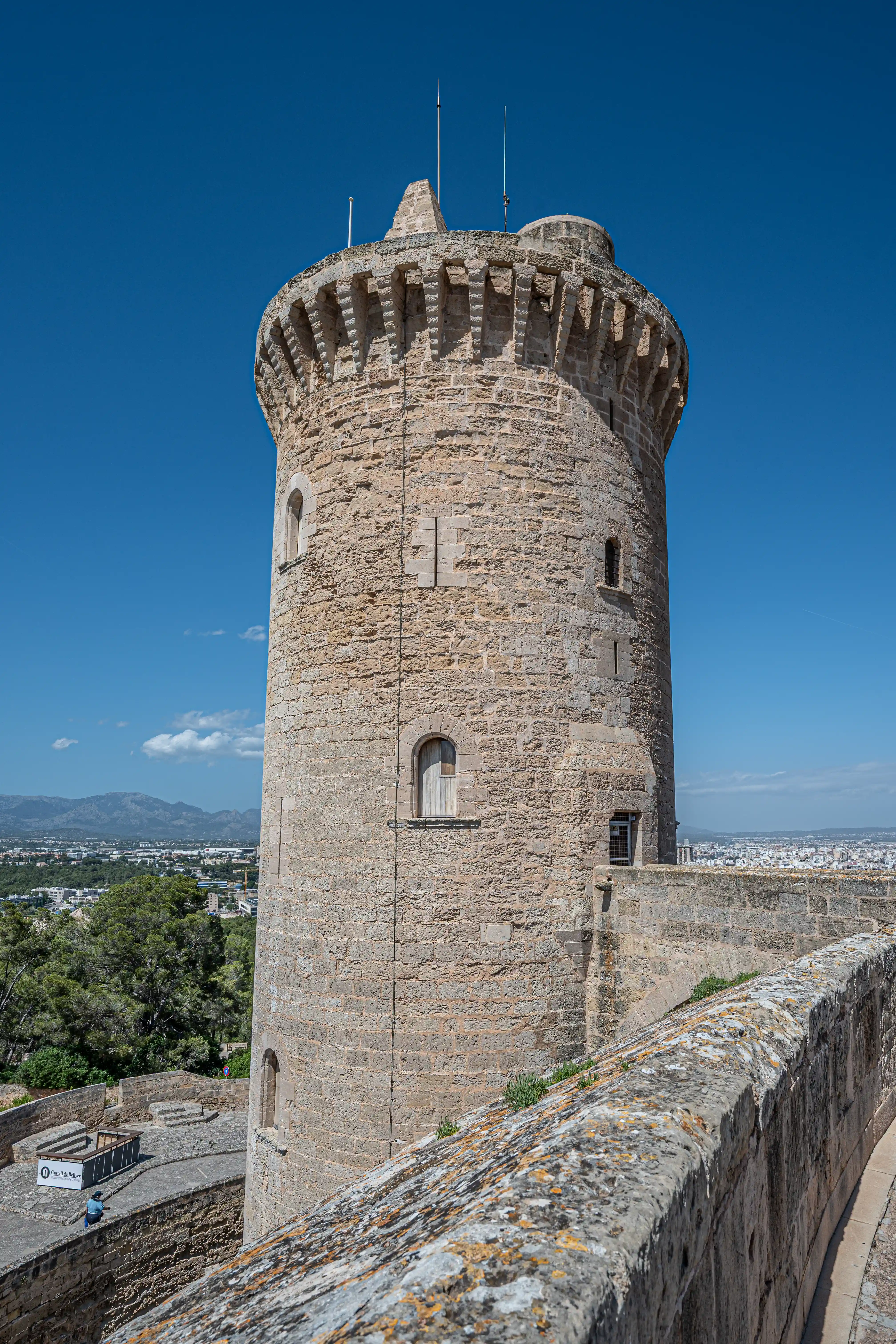A round, stone castle tower with crenellations on top, set against a bright blue sky. Small arched windows are visible on the tower's facade. A stone wall runs in the foreground, and a cityscape with mountains is in the background.