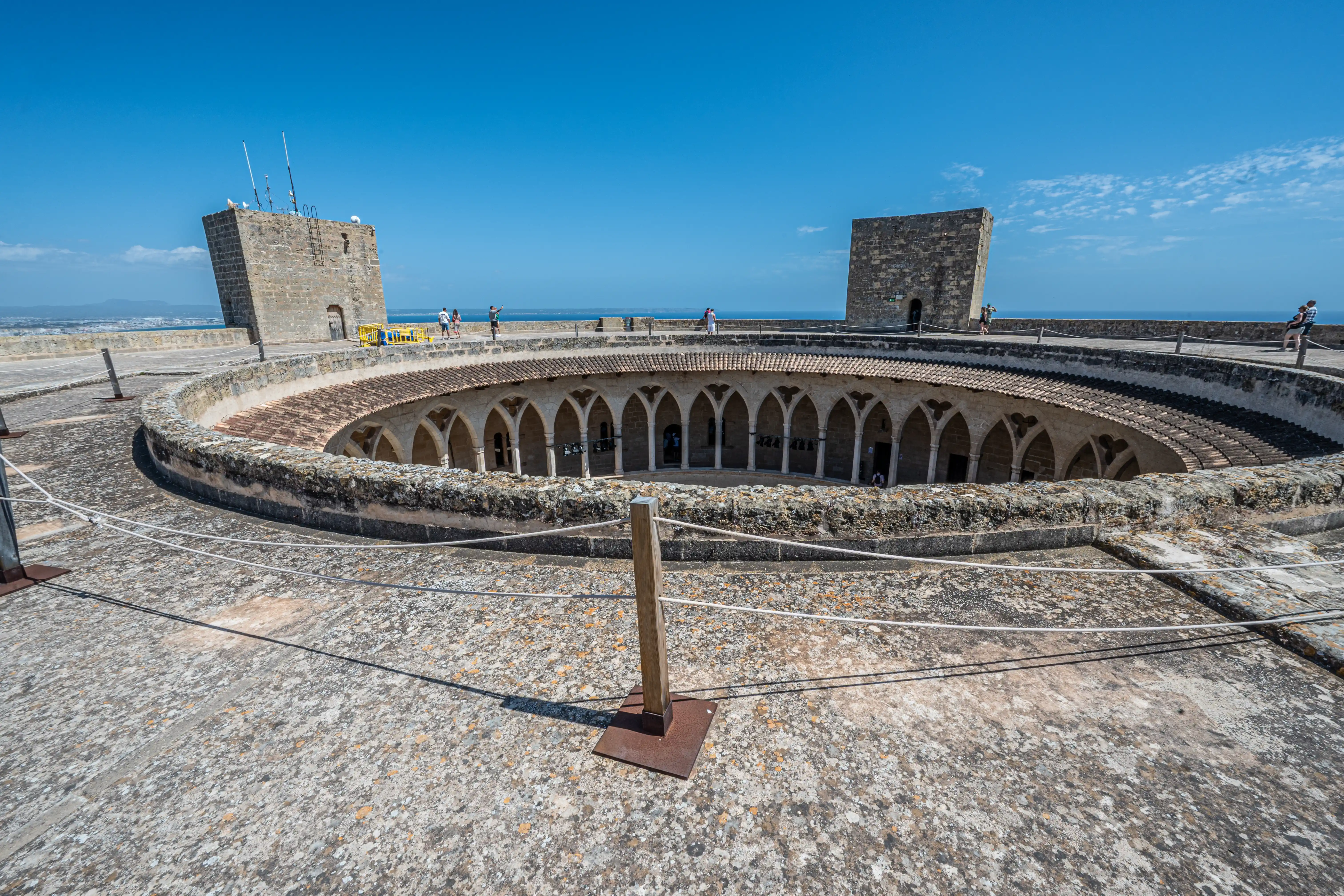 A circular courtyard within an ancient stone castle, showcasing a colonnade of arches and two imposing towers. The background is dominated by the calm blue sea and a bright, cloudless sky. A few visitors are visible walking along the castle's ramparts.