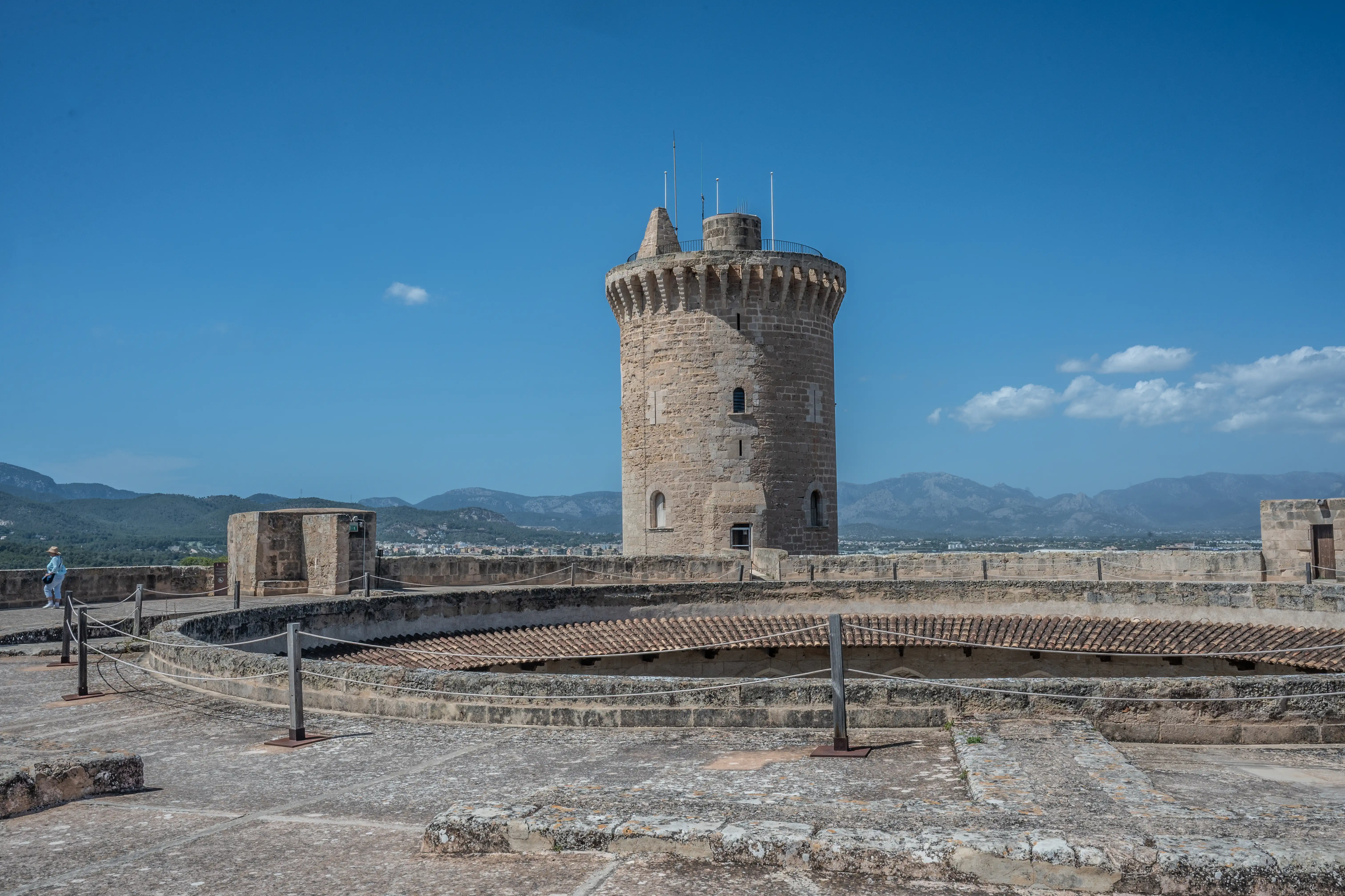 A stone castle tower with crenellations and windows, set against a bright blue sky. In the foreground is a stone courtyard with rope barriers, and in the distance are mountains and a town. A person walks along the ramparts in the left of the frame.